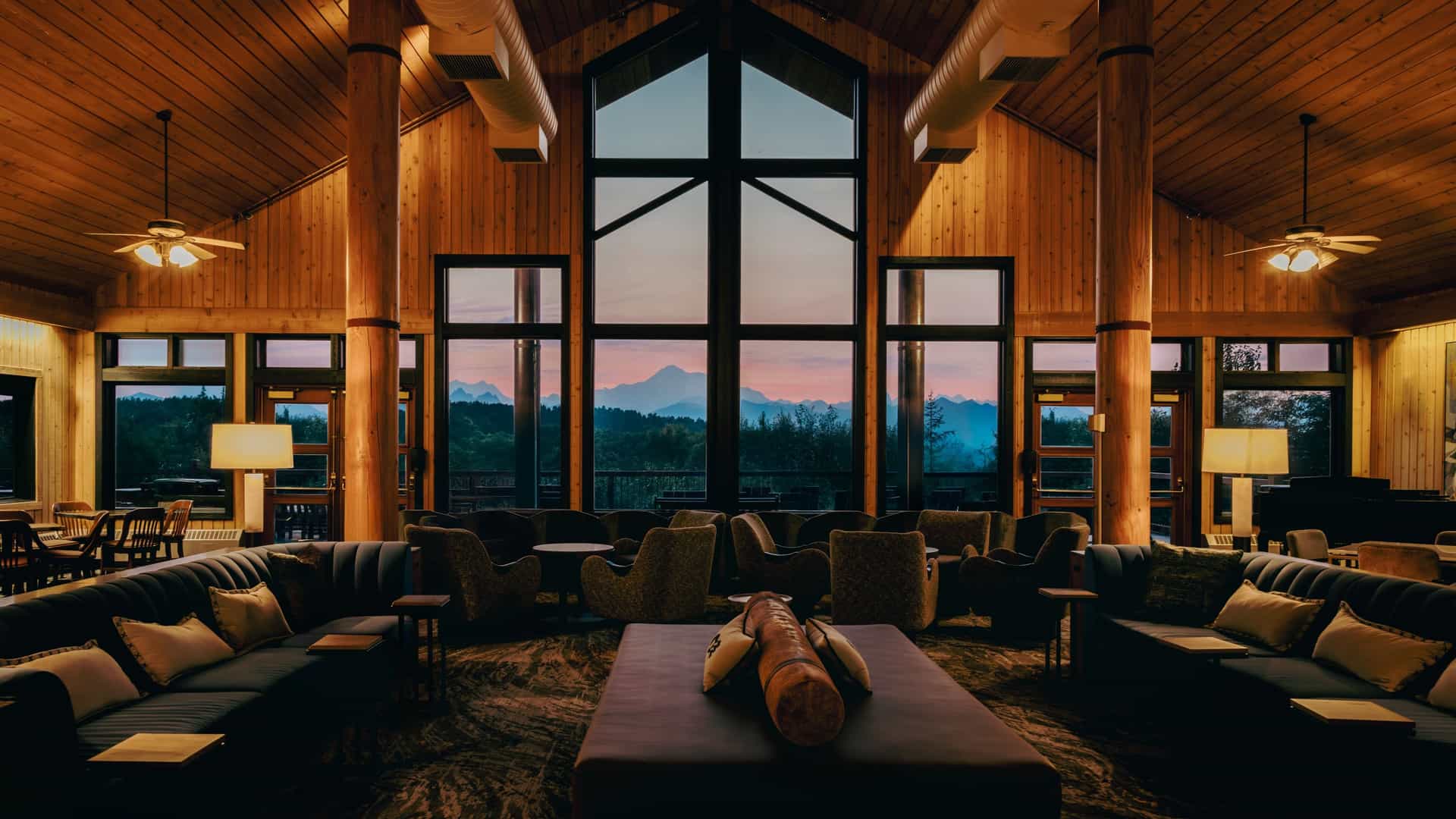 Cozy lodge great room with vaulted wood ceilings, large windows overlooking distant mountains at sunset, and comfortable sofas and chairs arranged for relaxing.