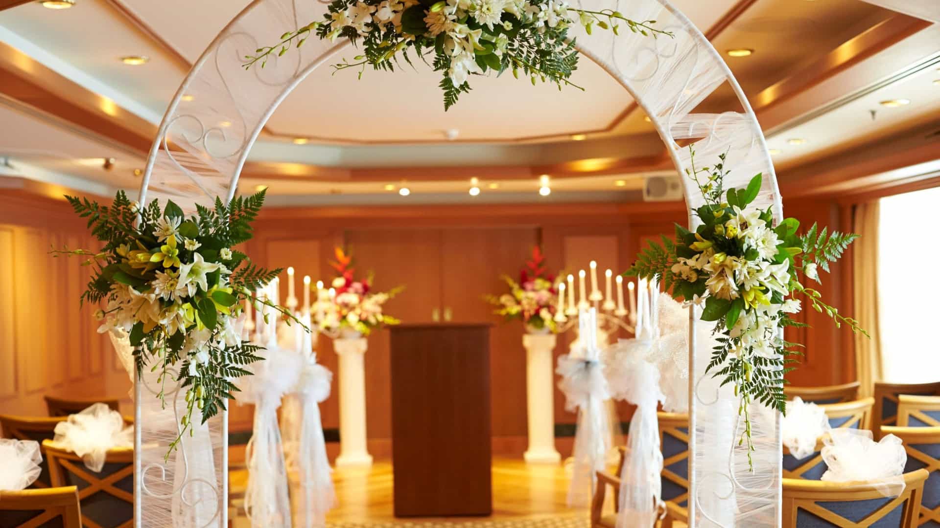 An elegant indoor wedding ceremony venue setup featuring a white decorative arch adorned with floral arrangements, leading toward a wooden podium, lit candelabras, and tulle-wrapped chairs.