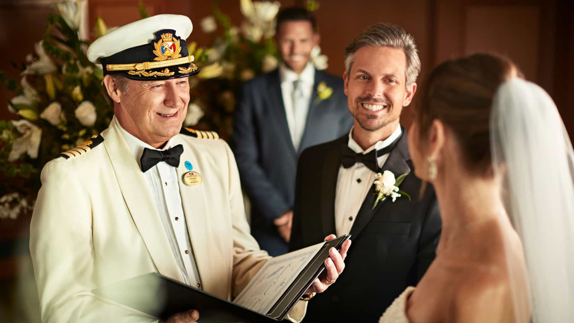 A Princess cruise ship captain in a white uniform officiates a wedding ceremony for a smiling groom in a black tuxedo and a bride with a veil.