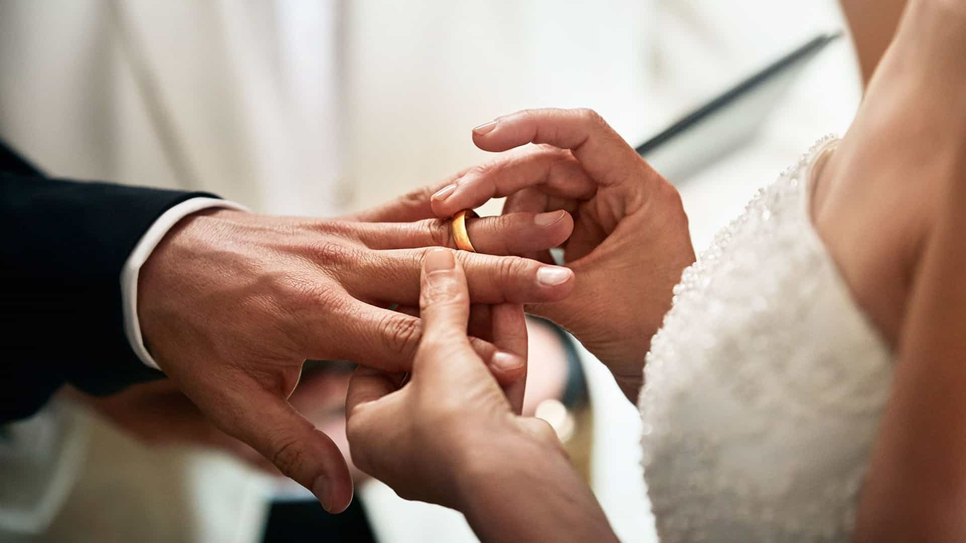 A close-up view of a bride placing a gold wedding band onto the groom's ring finger during their marriage ceremony.