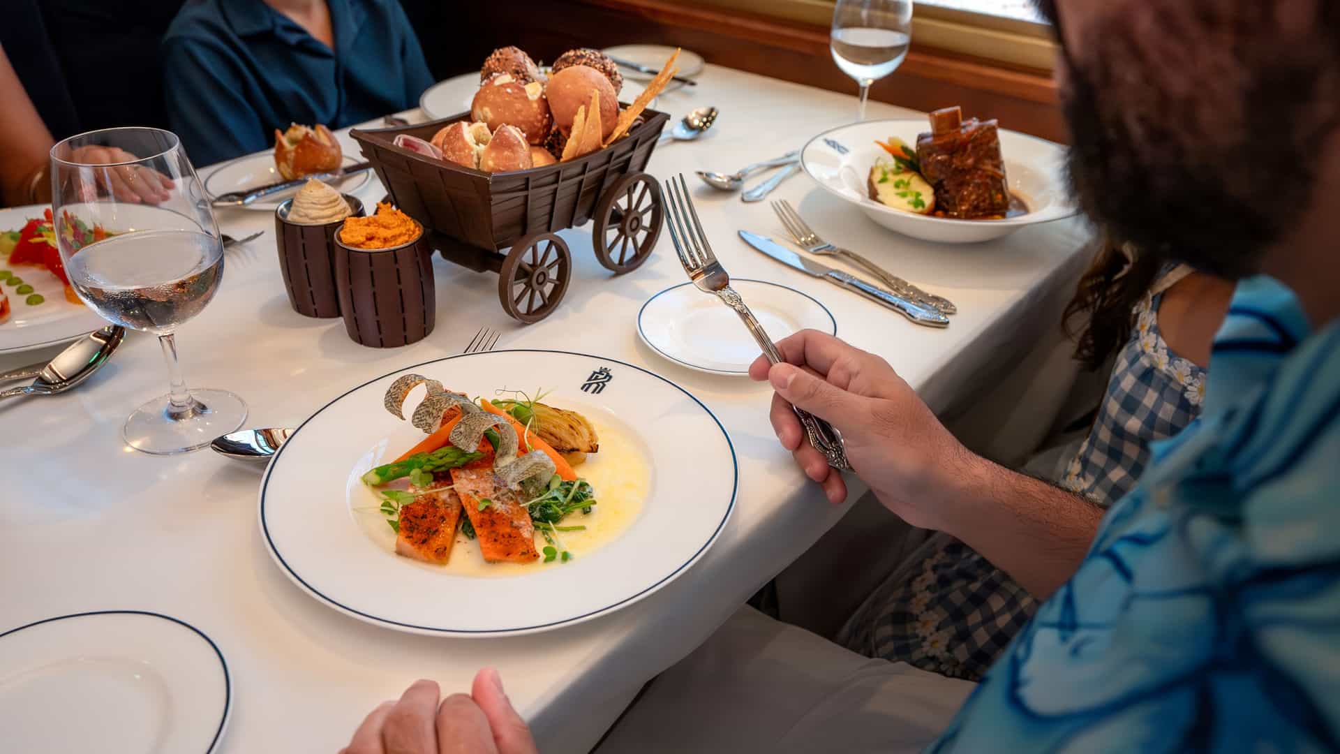 Over the shoulder shot of a man's meal aboard the Royal Railway aboard Utopia of the Seas. He is accompanied by his junior child to the right. They have a railway inspired bread basket in front of them.