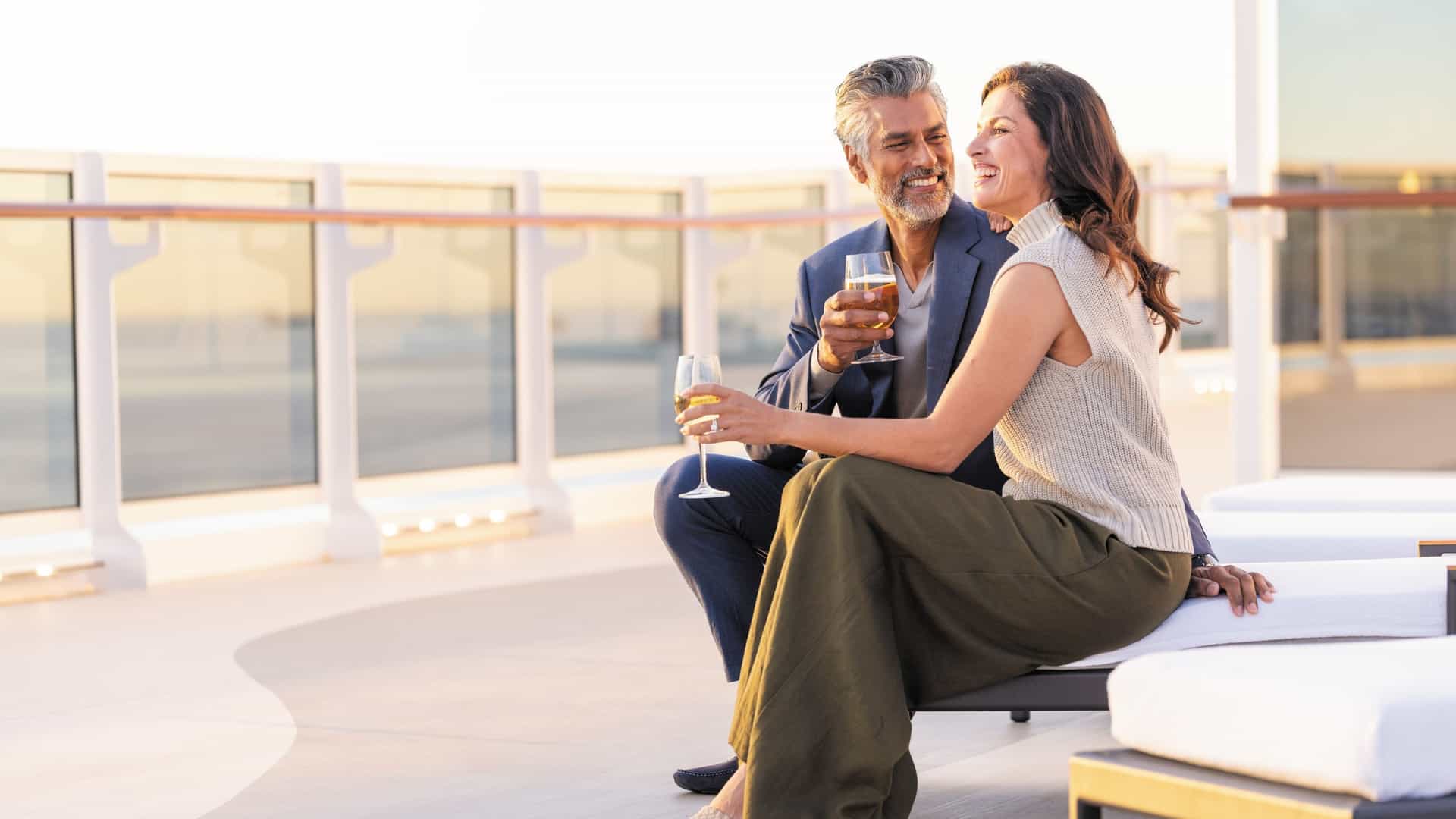 A smiling, formally dressed couple on the Regent Seven Seas Grandeur ship is sitting on lounge chairs on a balcony, clinking glasses for a toast.