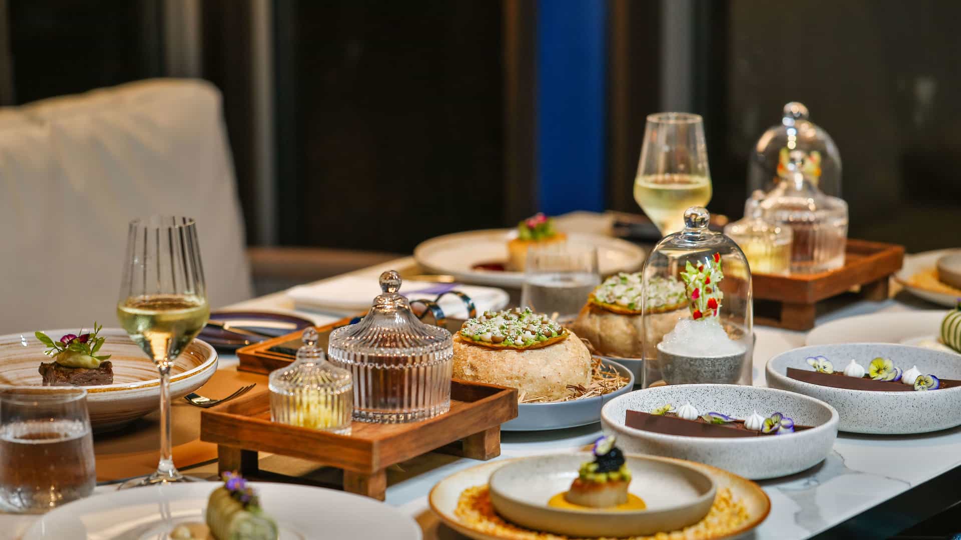 A beautifully set table for a gourmet dining experience at a fine restaurant. There are multiple plates of meticulously prepared food, each presented with elegant and artistic flair. The table is set with various glasses and fine cutlery, and the food includes miniature dishes under glass cloches. The scene captures the high-end, exclusive experience associated with culinary repositioning.
