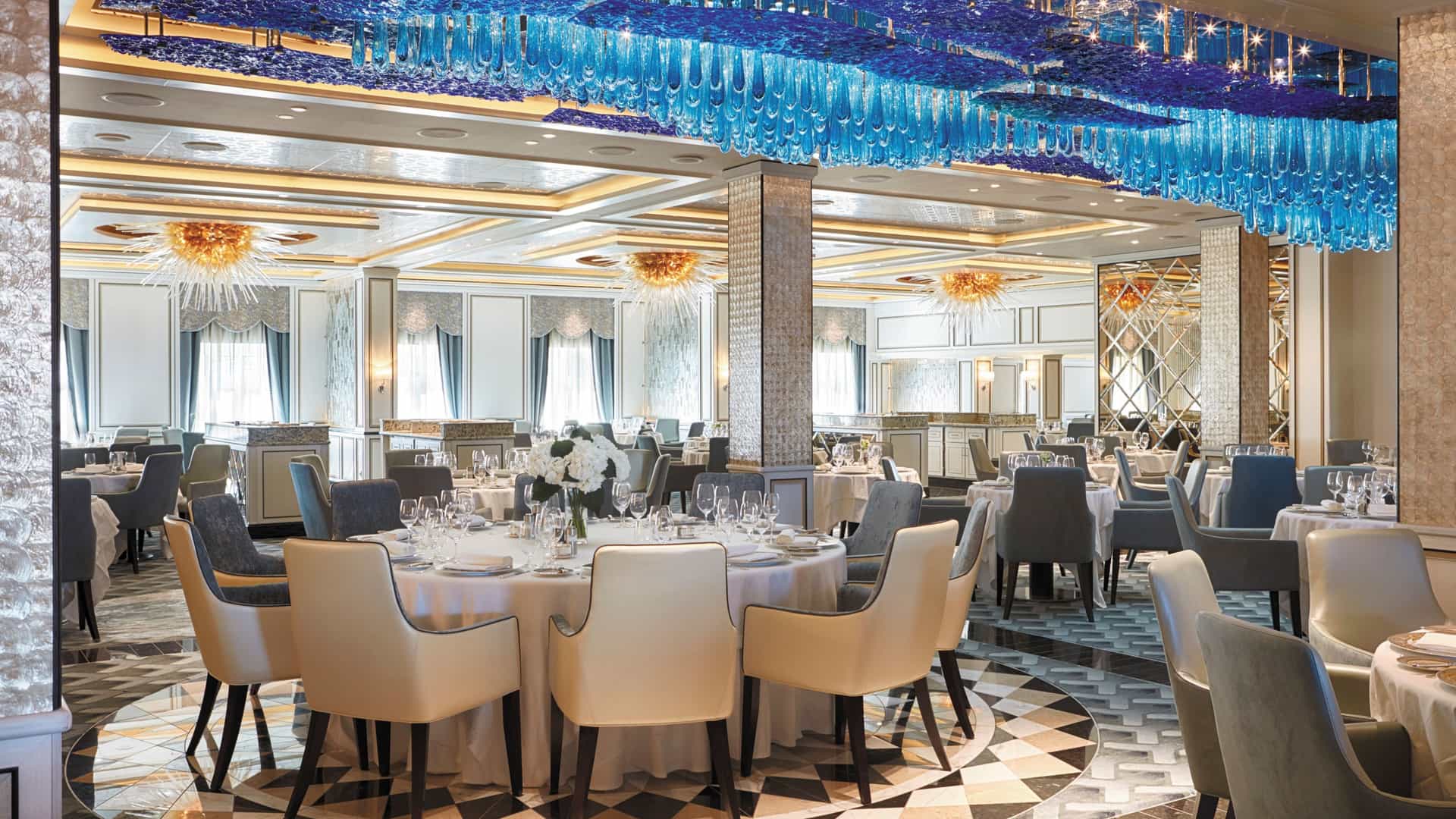 A wide shot image of the Compass Rose, an elegant dining venue aboard Regent Seven Seas. Showcasing a blue glass oceanic-like chandelier.