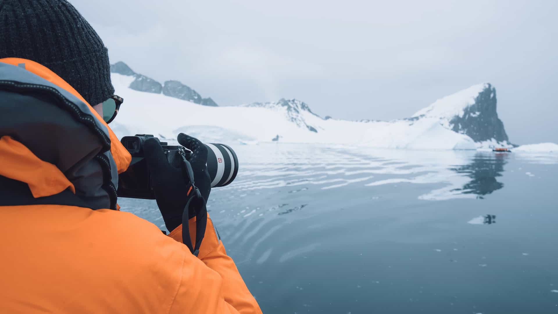 A person in a bright orange expedition parka and black beanie holds a professional camera with a telephoto lens, photographing a massive iceberg and snowy mountains in the Antarctic from the water.