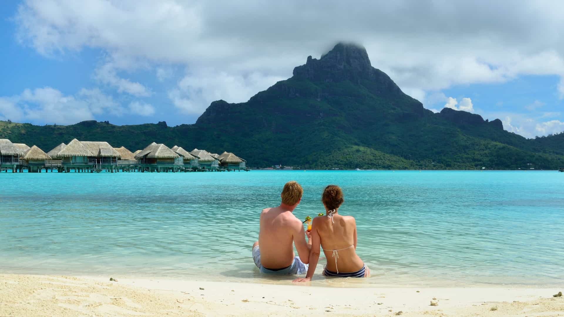 A couple relaxing on a pristine beach in Tahiti, French Polynesia, with overwater bungalows and a volcanic mountain in the background, a romantic destination for a luxury Seabourn cruise.