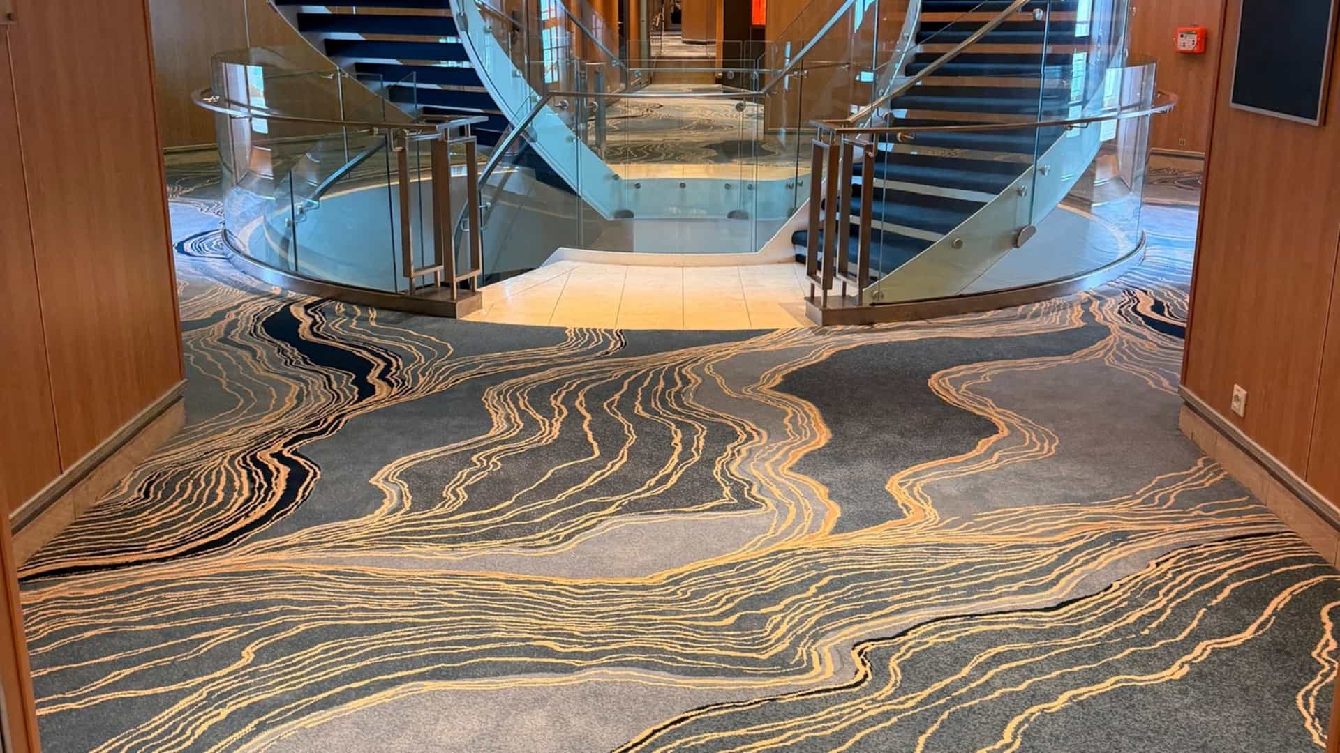 A view of the newly updated hallway carpet featuring blue and gold abstract wave patterns leading toward a curved grand staircase onboard the Seabourn Quest.