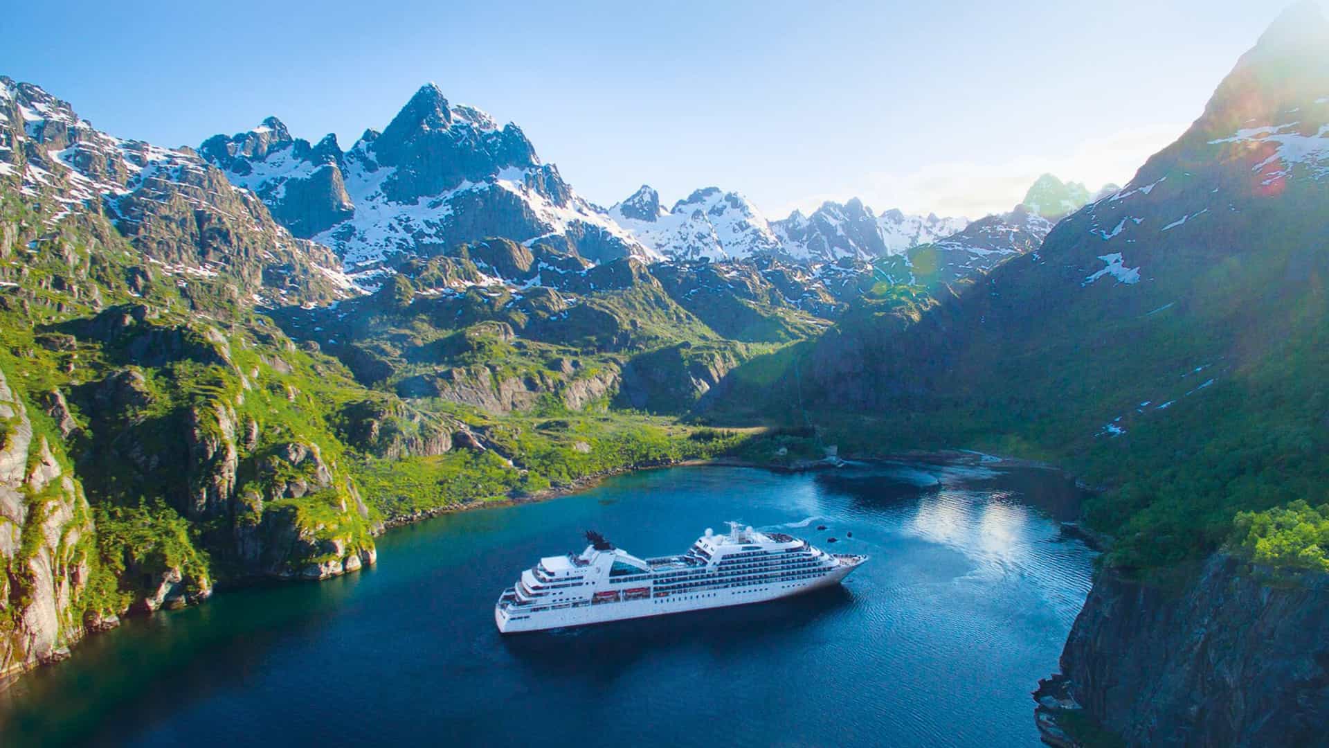 The Seabourn cruise ship navigates the calm, deep blue waters of a fjord, surrounded by majestic snow-capped mountains, indicative of a world cruise adventure.