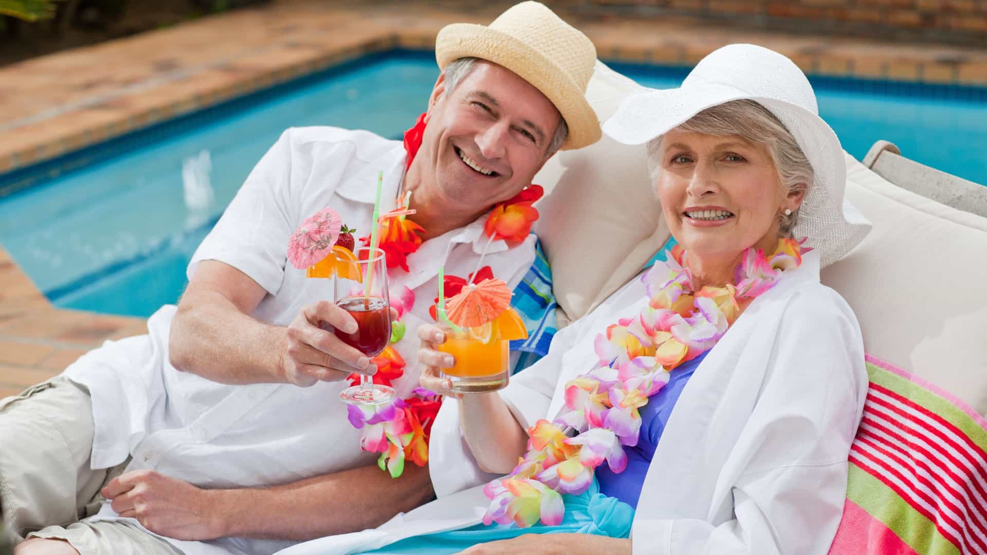A happy older couple sits by the pool, sipping drinks and enjoying a sunny day together on a cruise.