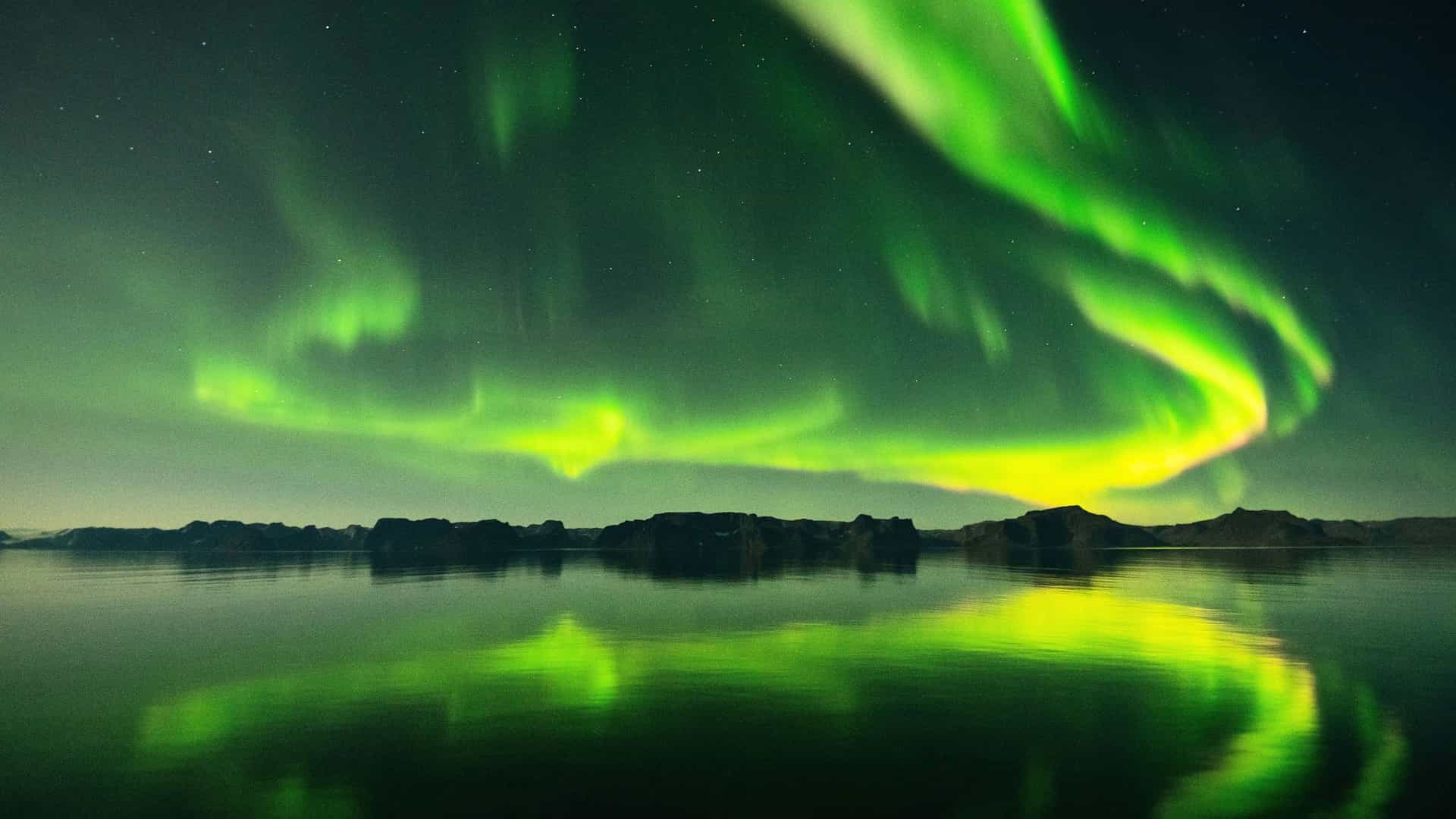 A vibrant green Northern Lights (Aurora Borealis) display illuminates the night sky and reflects on the water during a Silversea cruise in the Arctic Circle.