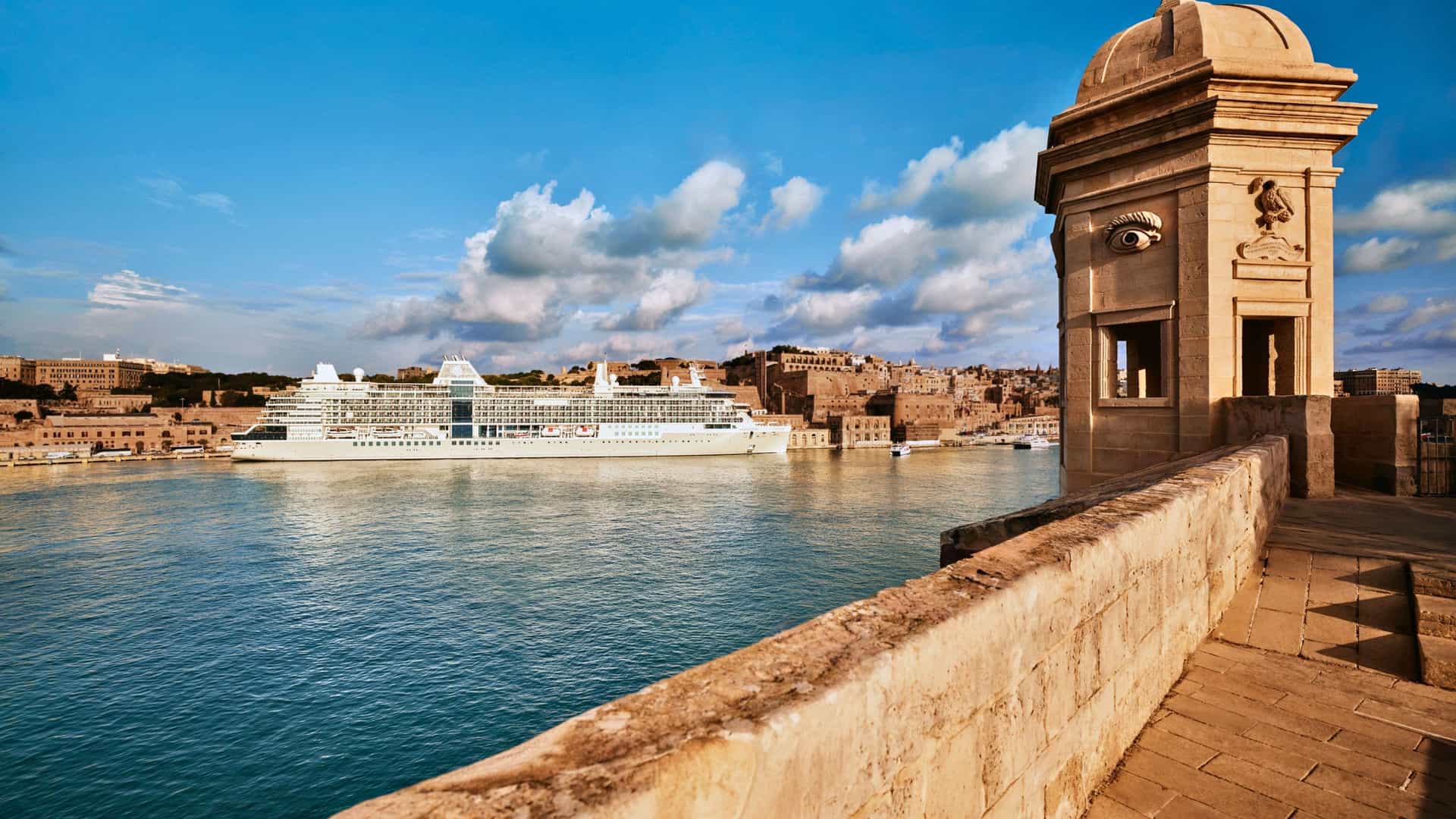 A Silversea cruise ship docked in the historic harbor of Valletta, Malta, with a classic stone watchtower in the foreground.
