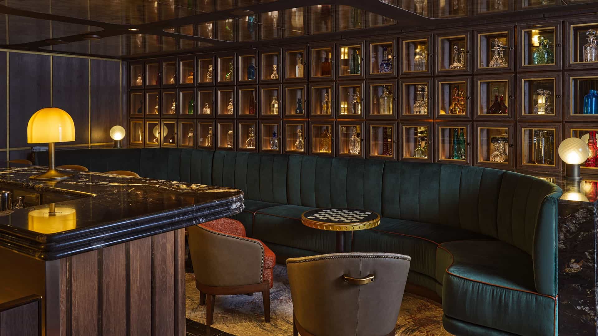 A sophisticated bar features a curved green velvet banquette, a dark marble counter with warm lamp lighting, and a wall of illuminated grid shelving displaying various glass decanters.