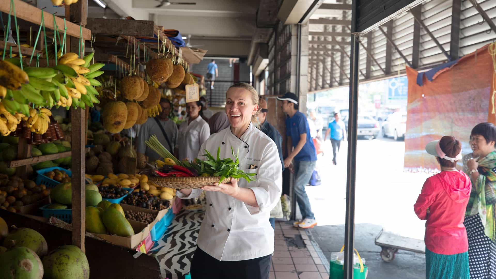 A smiling chef in a white uniform holds a woven tray of fresh greens and herbs while browsing a bustling local market filled with tropical fruits and produce.