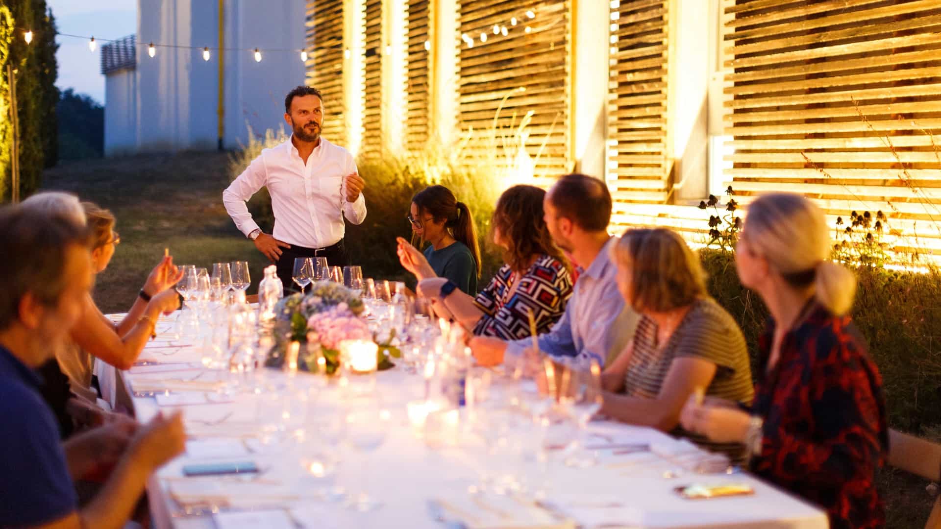 A man in a white button-down shirt stands and addresses a group of guests seated along a long, candlelit outdoor dining table at dusk.