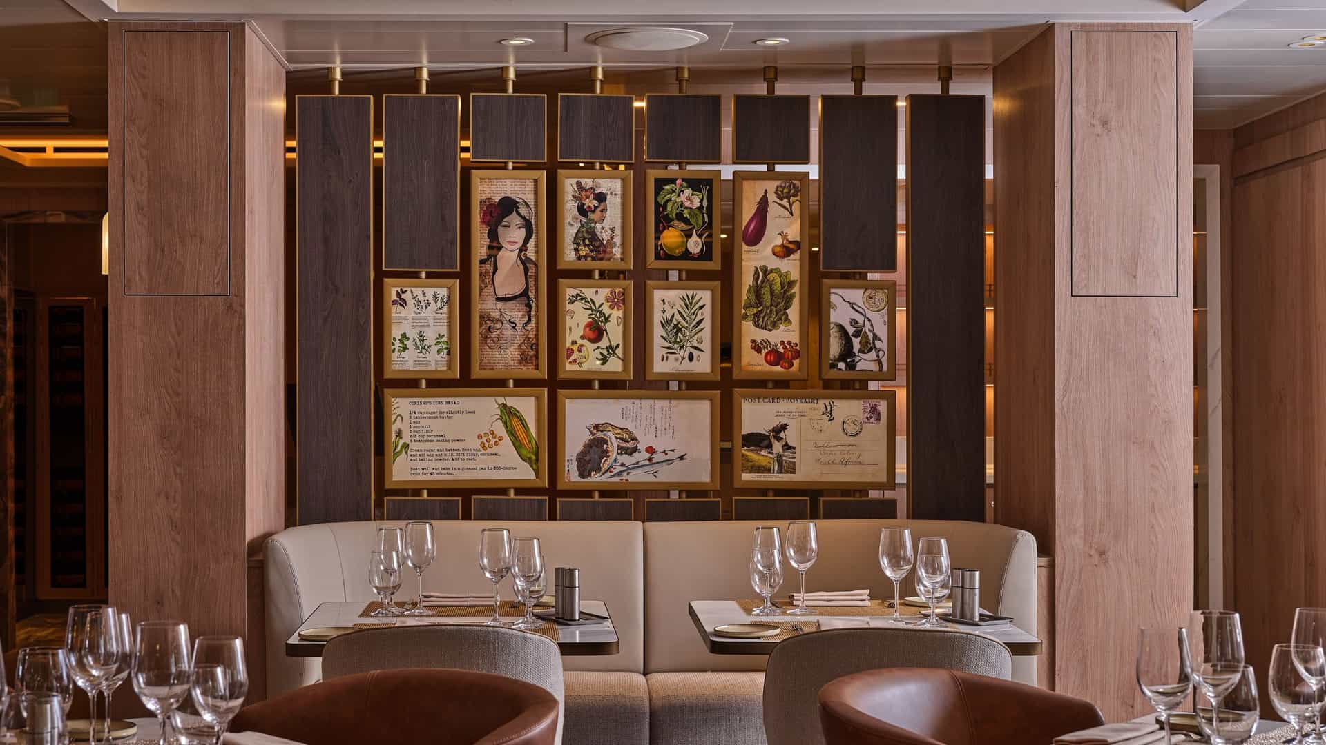 An elegant restaurant interior featuring a curved beige banquette booth beneath a decorative wall partition displaying framed botanical and culinary art prints.