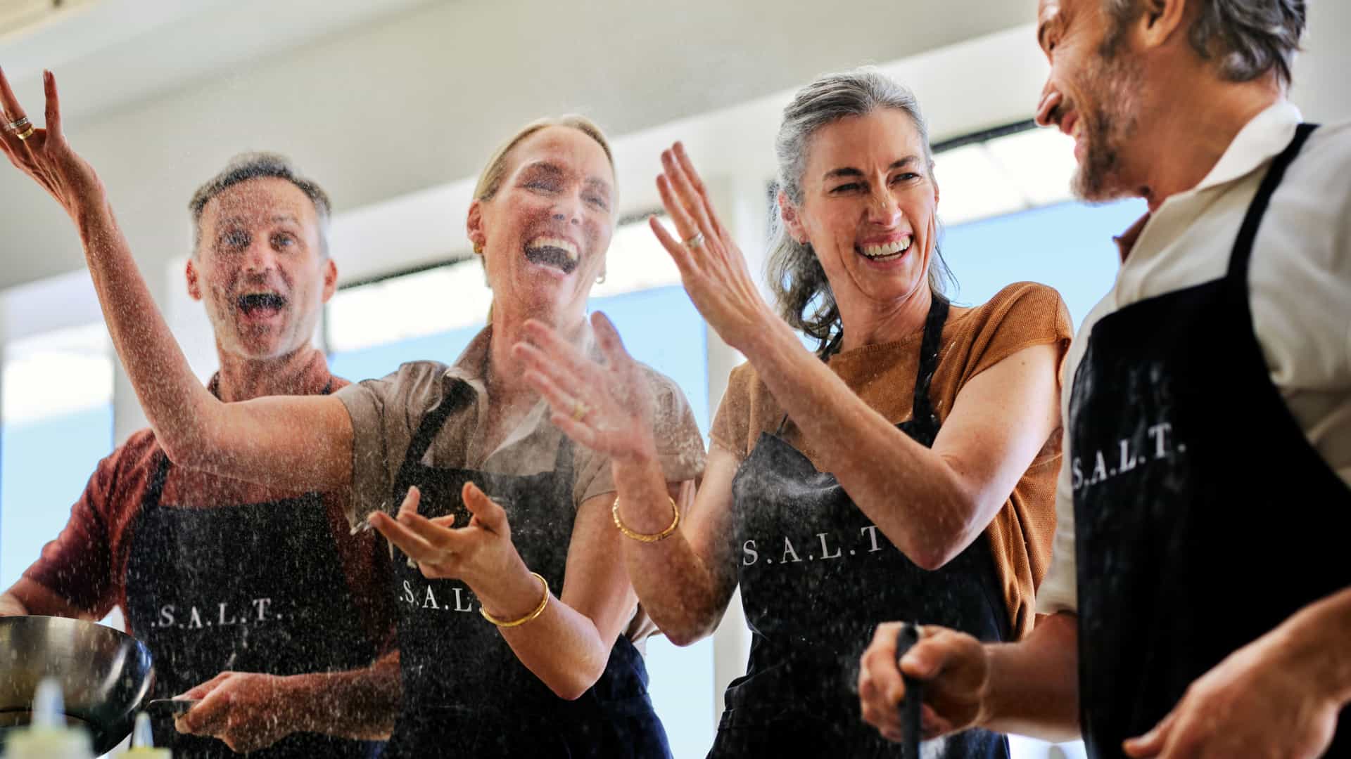 A group of 2 women and 2 men in aprons at the Salt Lab on Silversea's Silver Ray, laughing and enjoying themselves as they work through a culinary workshop at sea.