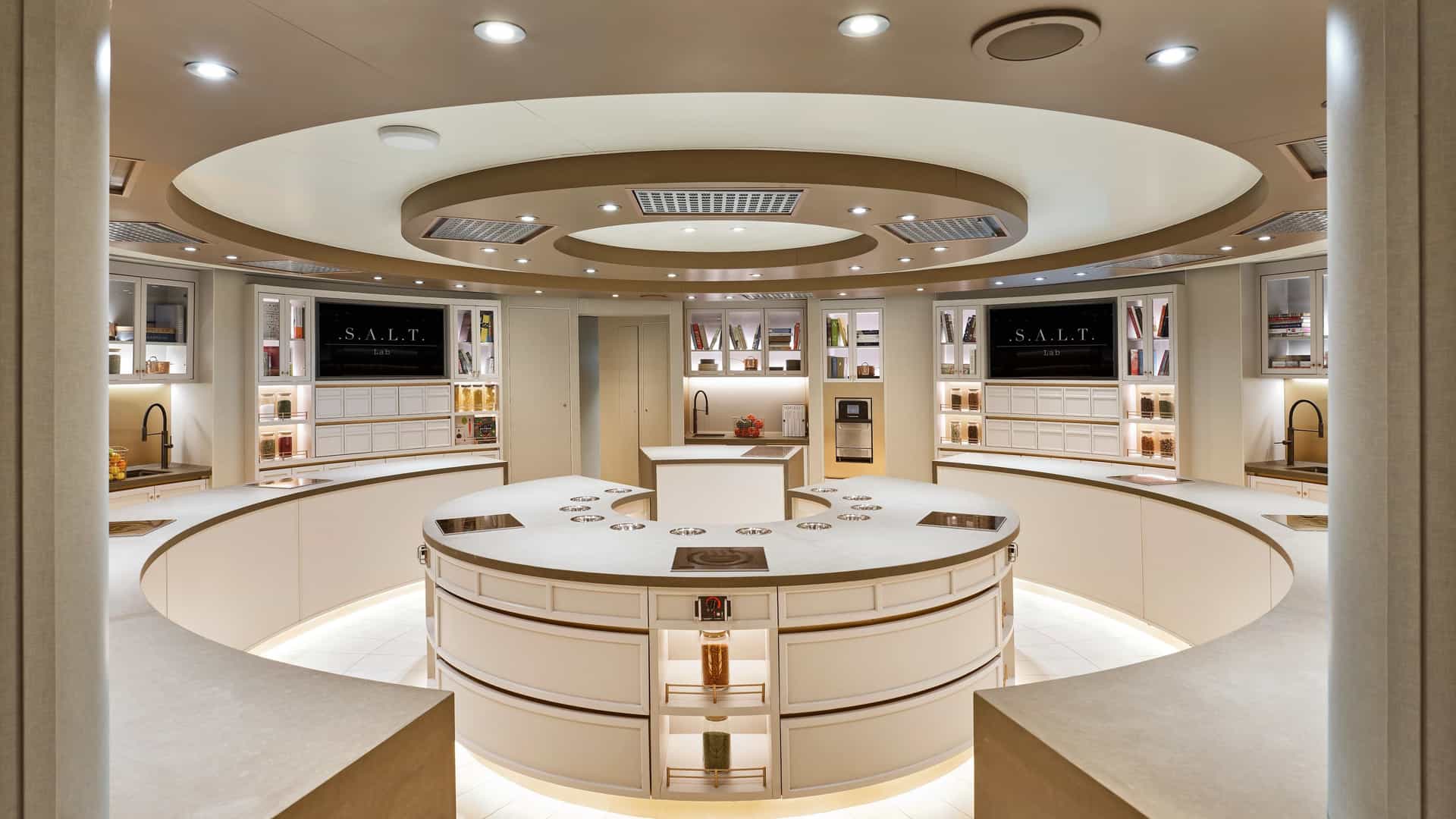 A modern culinary lab features a large circular central island surrounded by curved white counters, illuminated shelving, and a multi-tiered circular ceiling with recessed lighting.