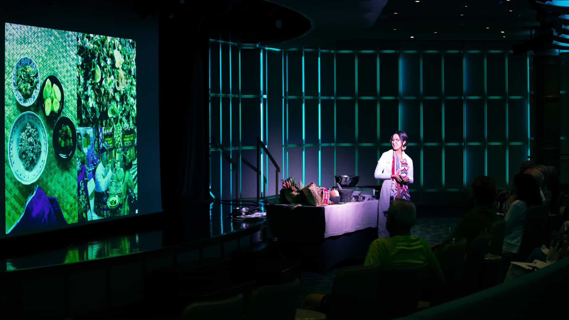 A speaker stands on a lit stage presenting to an audience in a darkened theater, next to a large projection screen displaying culinary images against a modern teal-illuminated wall.