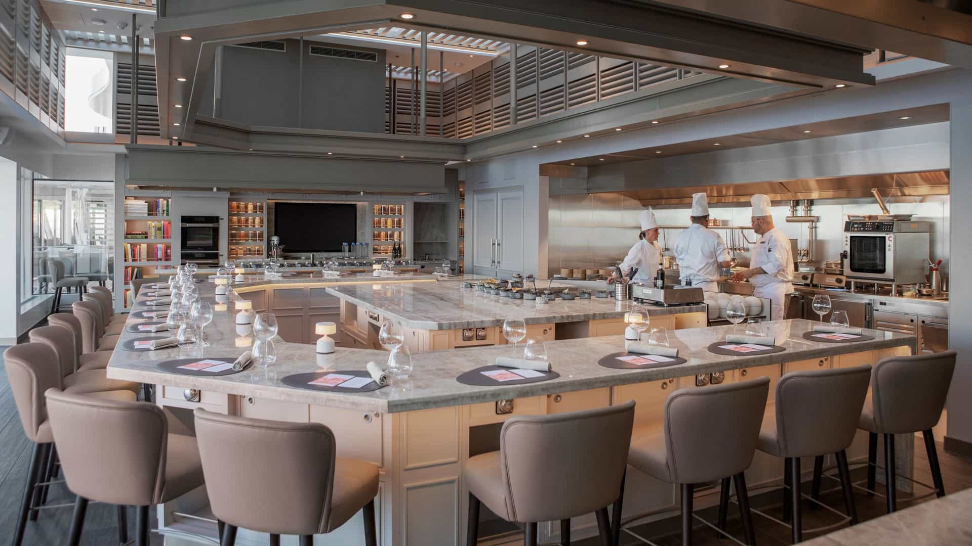 An elegant Chef's Table venue features a horseshoe-shaped marble dining counter with upholstered bar stools facing an open stainless-steel kitchen where chefs prepare meals.