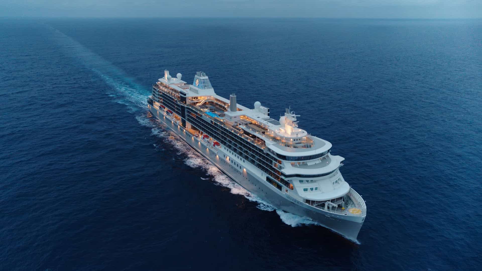 An aerial shot of Silversea's Silver Ray, sailing with cloudy skies and its evening exterior lights on.