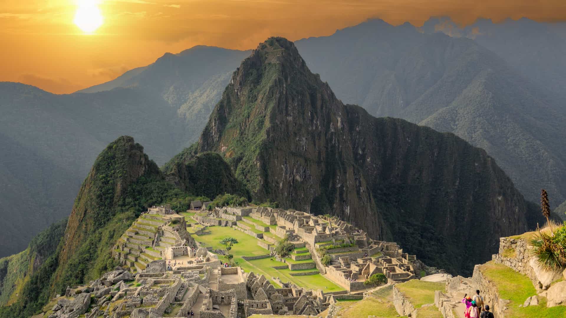 Hikers walk along a stone path overlooking the ancient stone ruins of Machu Picchu nestled among dramatic green Andes mountains beneath a glowing golden sunset sky.