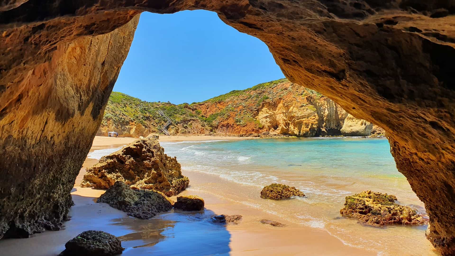 A rocky cave opening framing a Warrnambool beach with scattered shoreline rocks, clear turquoise water, and green‑covered cliffs under a bright blue sky.
