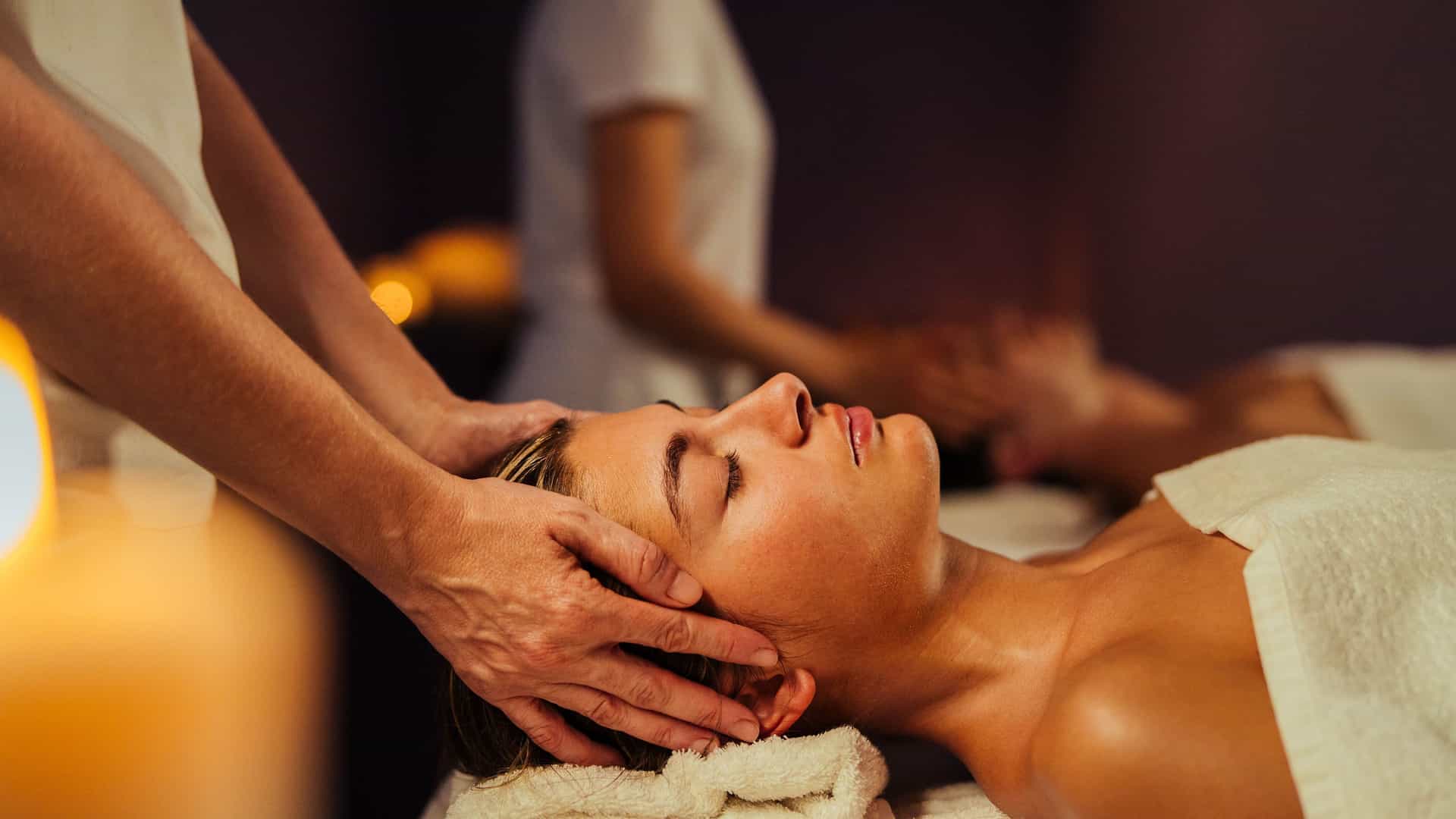 A serene scene aboard a cruise ship spa, where a woman lies on a massage table with her eyes closed. A masseuse's hands are gently positioned on the sides of her head, providing a relaxing head massage. The woman is wrapped in a white towel, and another person is visible in the background receiving a similar treatment. The warm, dim lighting and tranquil atmosphere convey the luxurious and relaxing amenities available during a trans-Atlantic cruise.