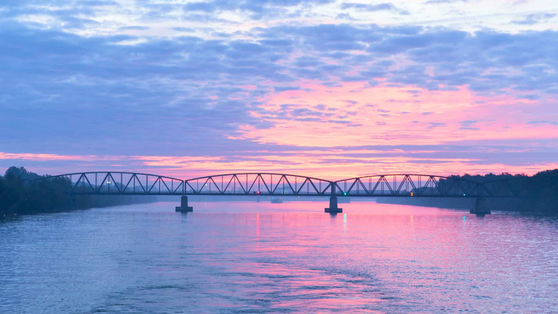A beautiful sunset over the Danube River, with a bridge silhouetted against a sky painted in shades of pink, purple, and orange, capturing a peaceful moment on a Uniworld river cruise.