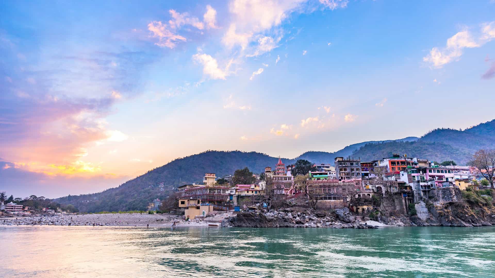 A scenic view of the Ganges River at sunset, with a vibrant riverside town nestled at the base of lush green hills, reflecting the unique cultural landscapes of a Uniworld river cruise in India.