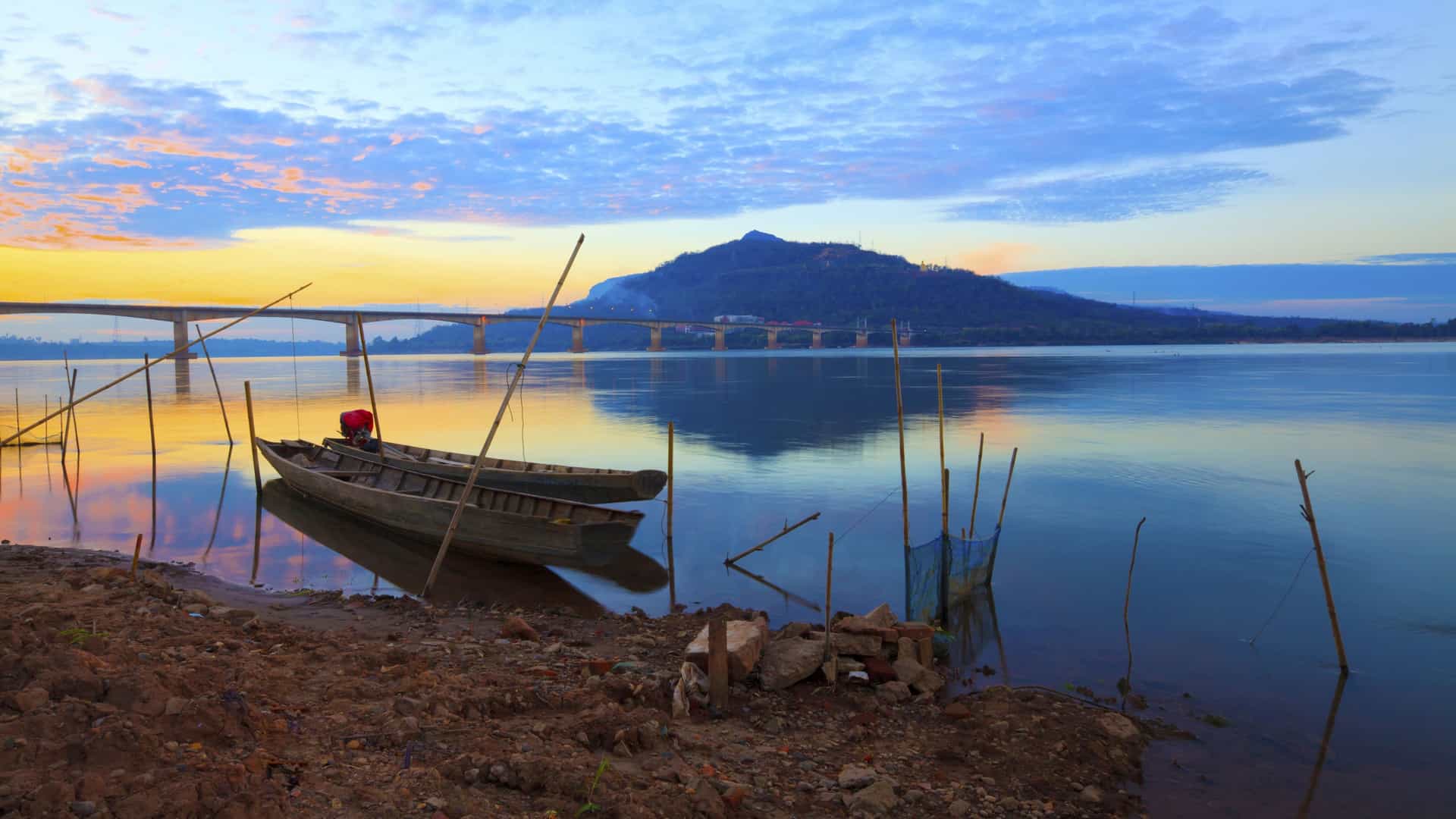 A stunning sunset over the Mekong River, with two small boats resting near the shore and a bridge in the distance, capturing the serene beauty of a Uniworld river cruise destination in Asia.