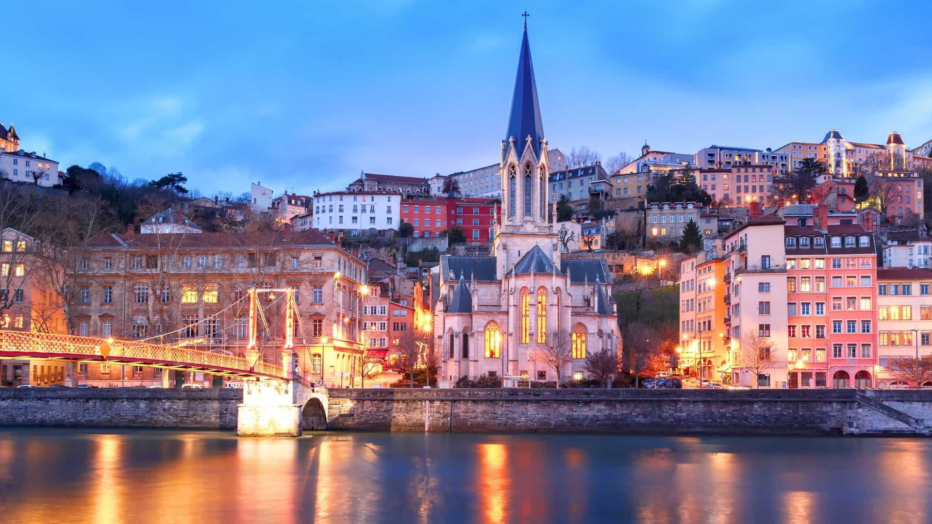 An evening view of the Saône River in Lyon, France, with a historic church and colorful buildings illuminated along the riverbank, a captivating sight on a Uniworld river cruise.
