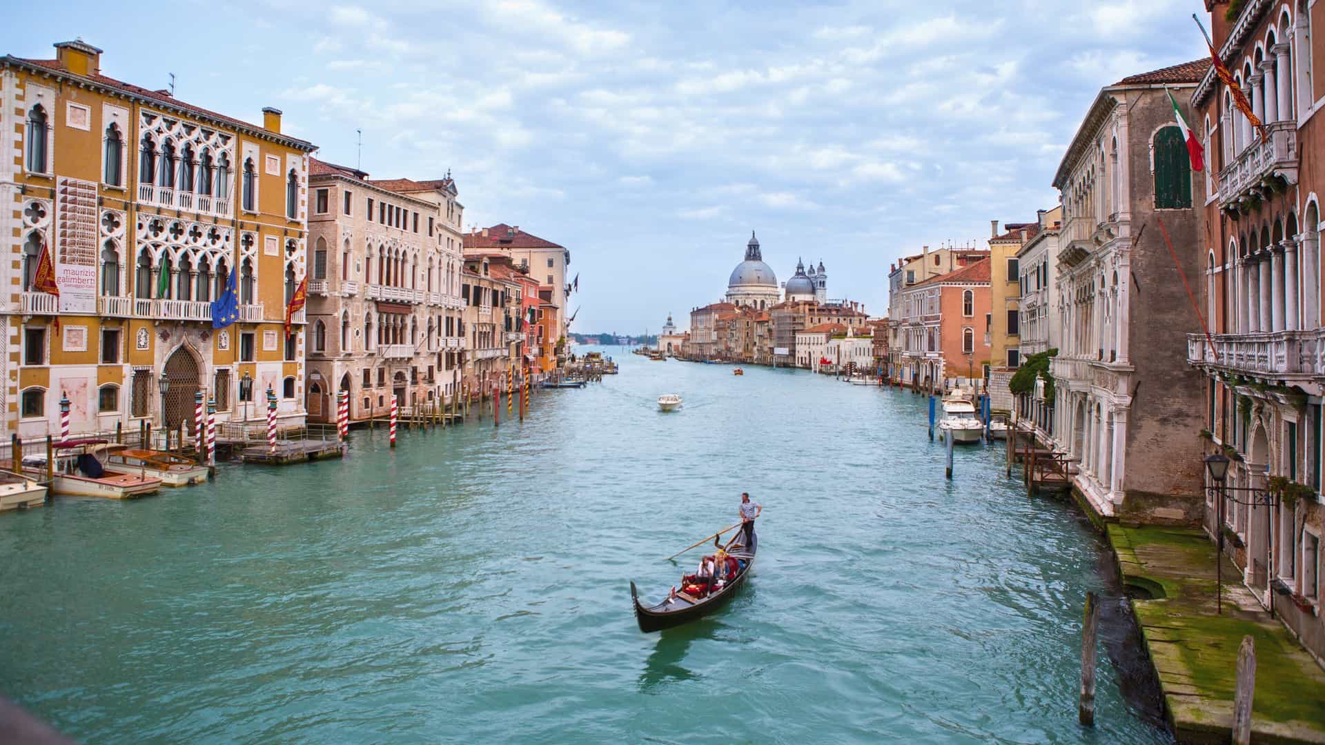A traditional gondola with people is seen on the Grand Canal in Venice, Italy, surrounded by historic buildings under a cloudy sky. The image captures the essence of a Uniworld river cruise destination in the Mediterranean.