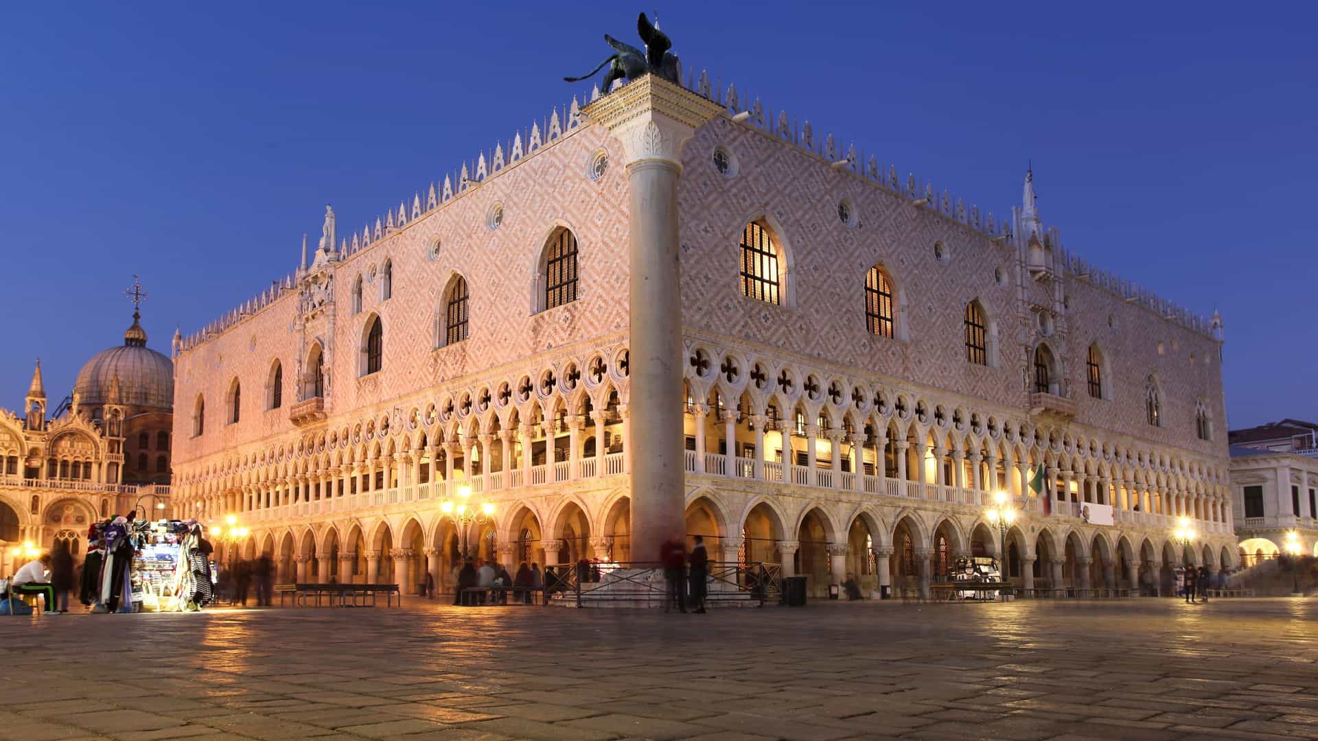 A night-time view of the Doge's Palace and St. Mark's Basilica at Piazza San Marco in Venice, Italy, with glowing lights illuminating the Gothic architecture under a deep blue twilight sky.