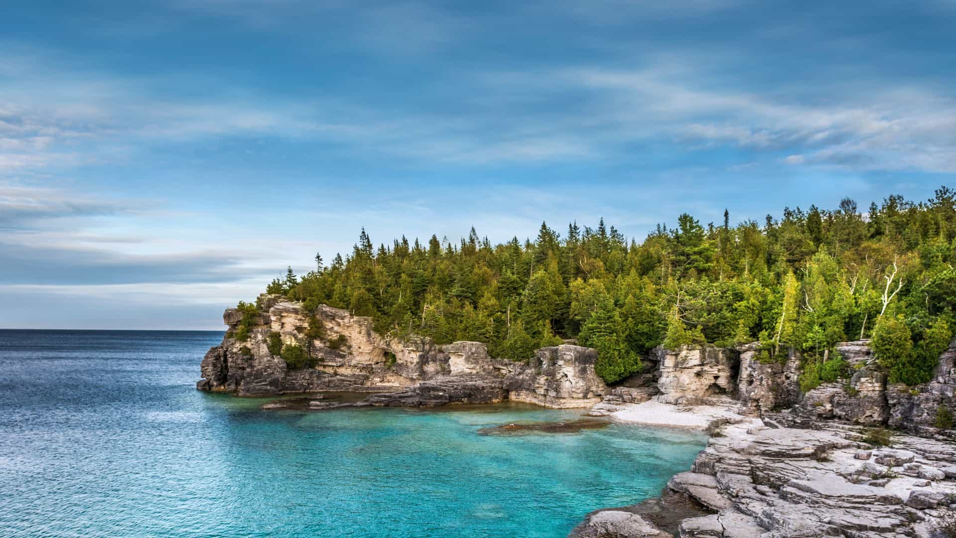 A stunning coastal scene in Ontario, Canada, featuring turquoise waters, rugged rocky cliffs, and dense evergreen forests under a partly cloudy sky—ideal for Viking Expedition Cruise exploration.