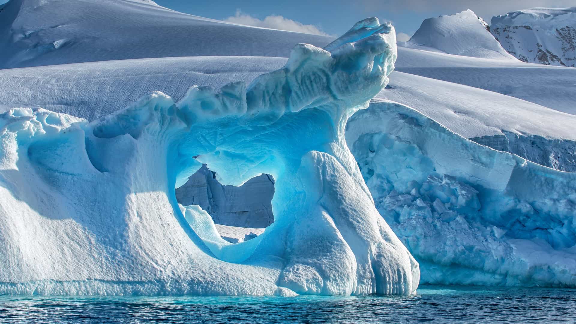 Icy landscape in Wilhelmina Bay, Antarctica, featuring a vivid blue iceberg with an arch formation, calm waters, and snow-covered mountains—an iconic view for Viking Expedition Cruise explorers.