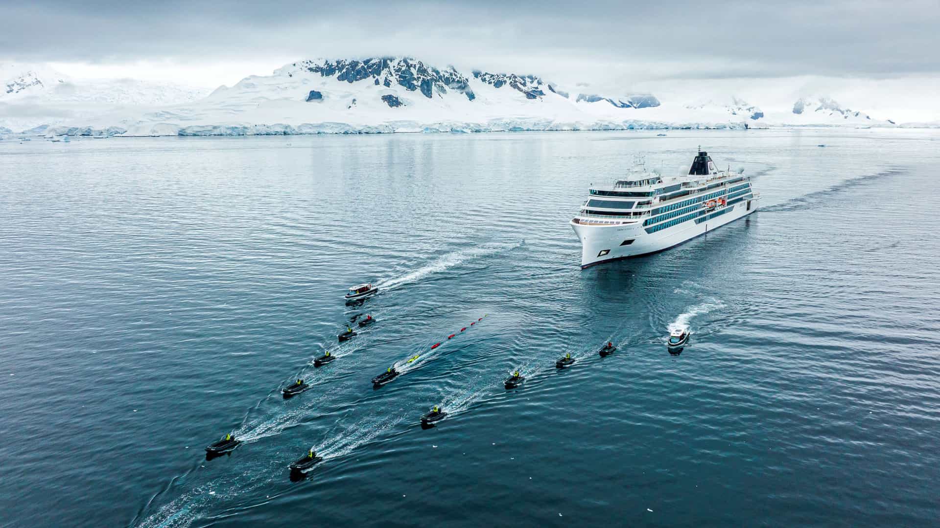 An aerial view shows a white Viking expedition cruise ship sailing in calm blue waters in Antarctica, trailed by a fleet of several small black Zodiac boats creating wakes. Snow-capped mountains and an overcast sky are visible in the background.