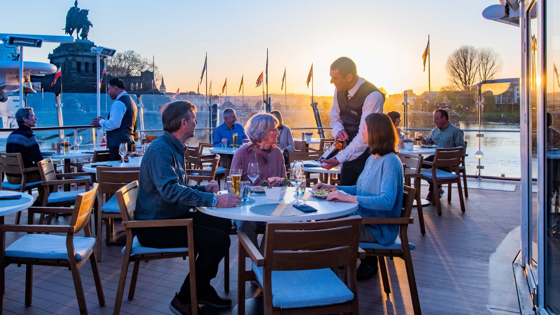 Guests enjoy outdoor dining and wine service on the Aquavit Terrace of a Viking river cruise ship during a scenic sunset in Koblenz, Germany.