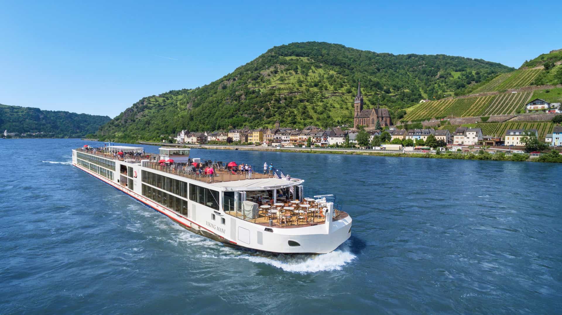 The Viking Mani river cruise ship glides along the Rhine River past a historic German hillside village and terraced vineyards in the Middle Rhine valley.
