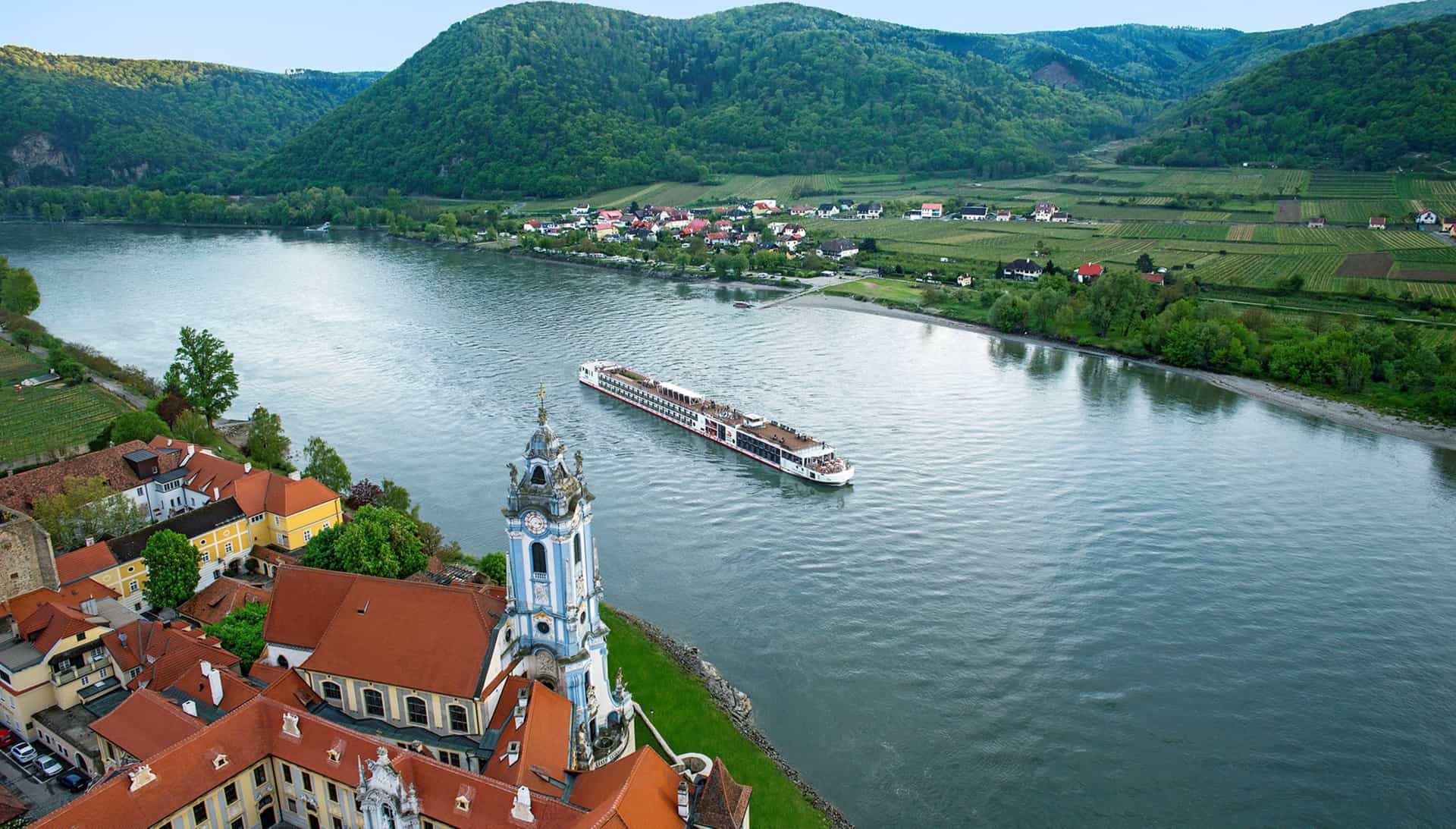 A Viking river ship sails down the Danube River, passing the blue and white bell tower of the Abbey of Durnstein in Austria, with vineyards and green hills in the background.