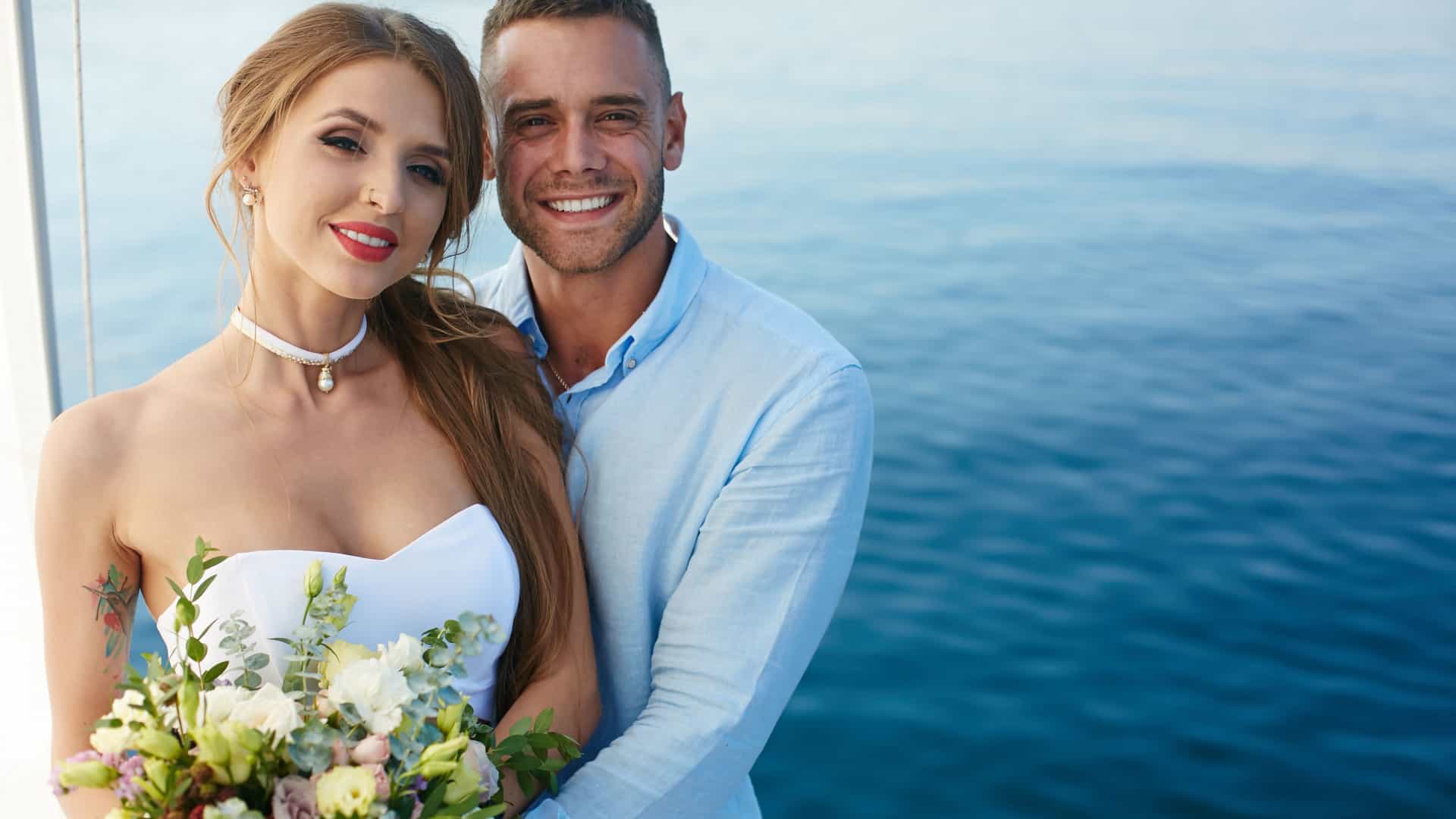 A bride and groom in formal wedding attire stand on a cruise ship, enjoying a romantic moment against a scenic backdrop.