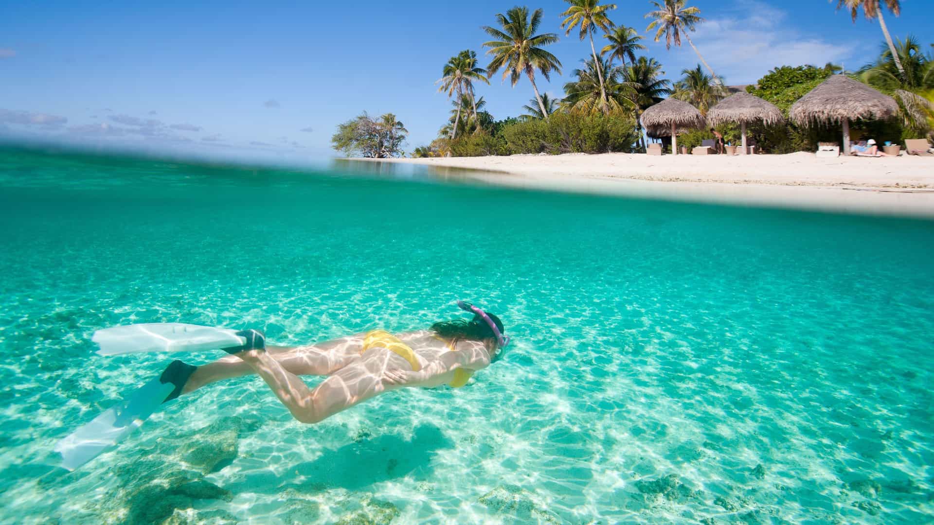 An over-under photograph showing a woman snorkeling in the clear, turquoise waters of the Western Caribbean. Below the surface, the woman is seen wearing a yellow bikini, a snorkel, and flippers as she swims over a sandy seabed with some rocks. Above the water, a pristine white-sand beach with palm trees and thatched-roof huts is visible under a clear, blue sky. The image perfectly captures a tropical beach vacation experience in the Caribbean.