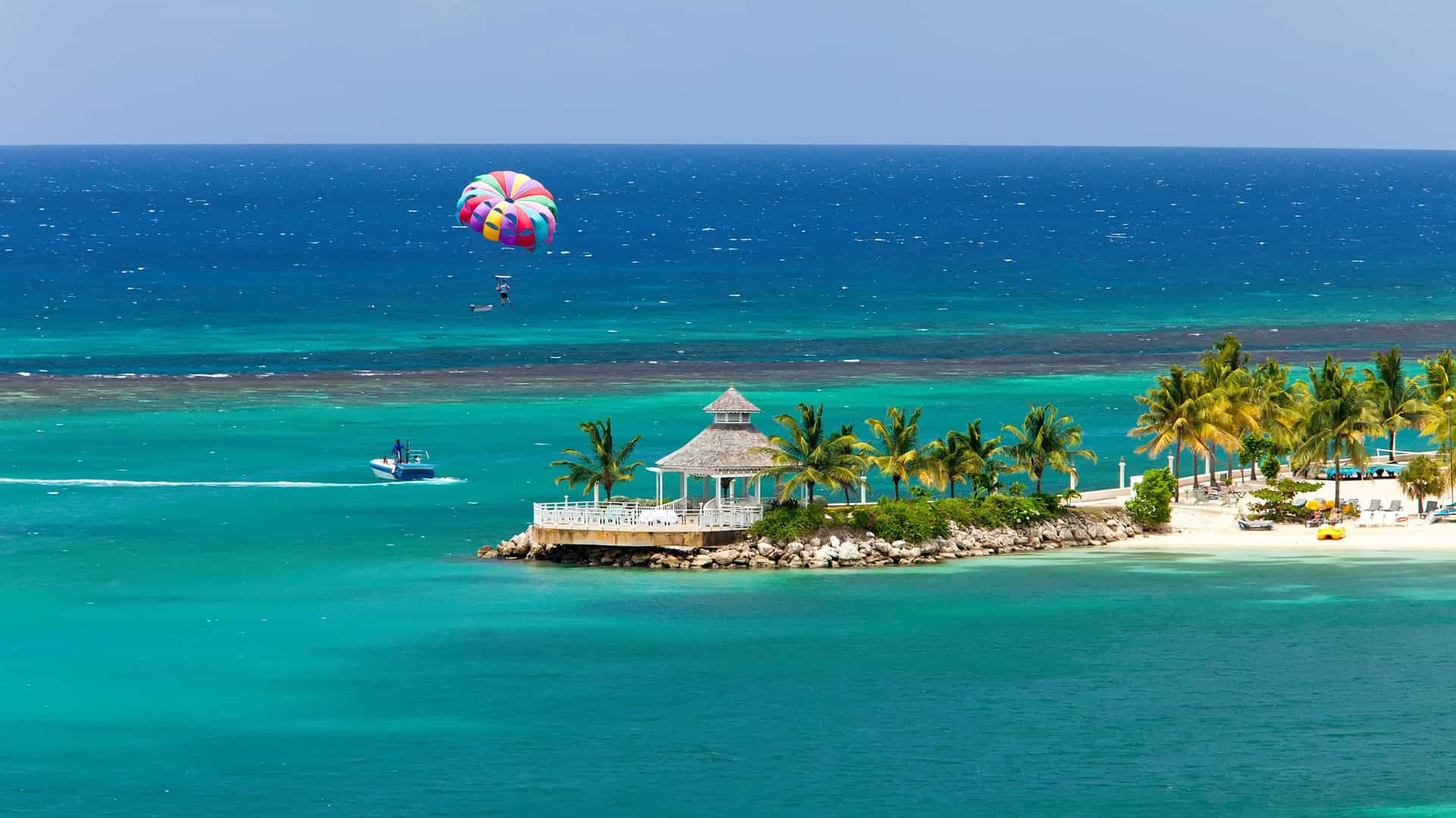 An aerial view of a person parasailing over the bright turquoise waters near the coast of Ocho Rios, Jamaica. The colorful parachute, with its red, blue, green, and yellow stripes, is high in the air, towed by a motorboat visible on the left. In the foreground, a small island with a white gazebo and palm trees sits in the clear water. The image captures a popular tourist activity and the stunning tropical scenery of the Western Caribbean.