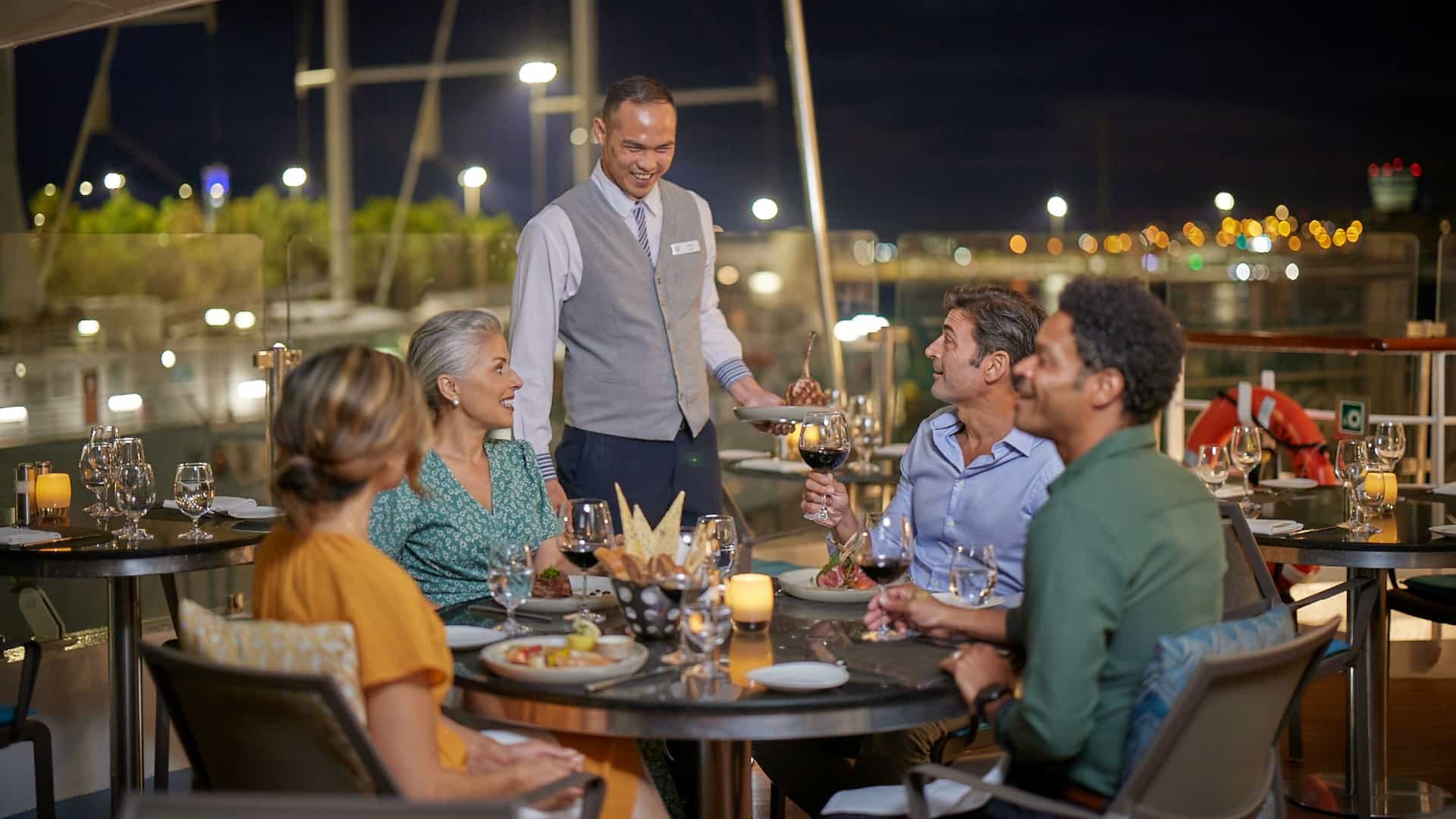 A smiling waiter serves a group of four friends enjoying an outdoor, candlelit dinner on the deck of a cruise ship at night, with city lights blurred in the background.