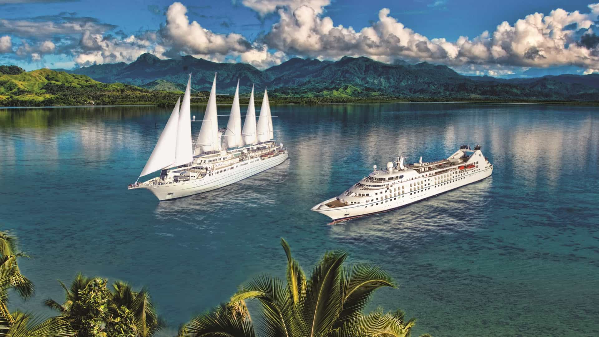 Two white Windstar cruise ships, one featuring prominent towering sails and the other a classic motor yacht design, sail through calm turquoise waters against a backdrop of lush, green tropical mountains.