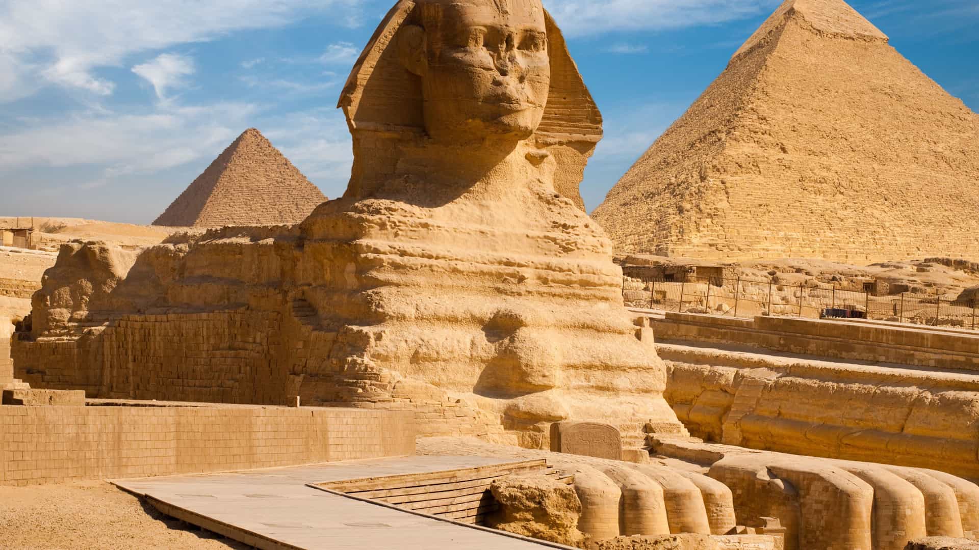 The Great Sphinx of Giza, a monumental limestone statue of a mythical creature with the body of a lion and the head of a human, sits majestically in the desert. In the background, two of the iconic pyramids of Giza rise against a clear blue sky with some white clouds. The entire scene is bathed in bright sunlight, highlighting the ancient, weathered stone of the Sphinx and the pyramids. The image captures the famous historical landmarks of Egypt.