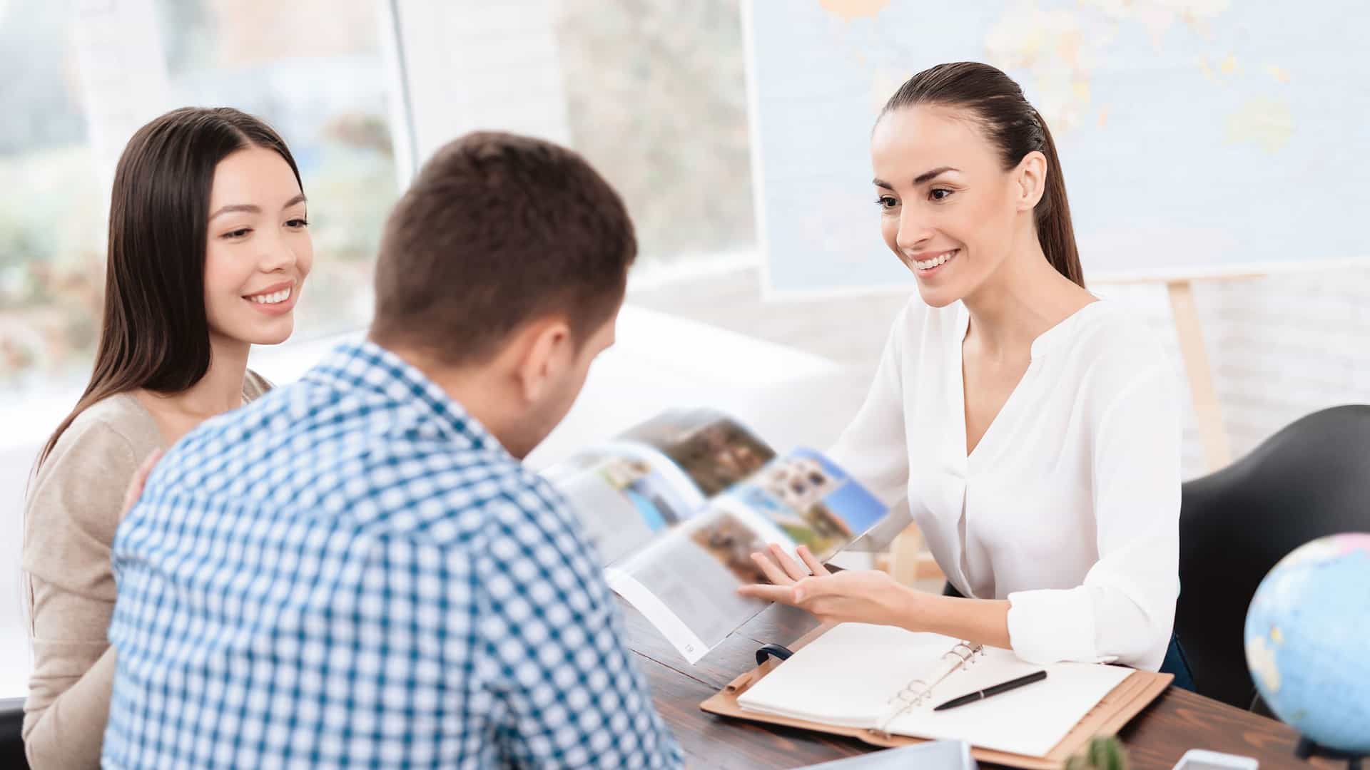 A friendly female travel agent sitting at a desk and smiling as she shows a brochure to a young couple. The couple, a man and a woman, are looking intently at the brochure with happy expressions on their faces, suggesting they are planning a vacation. The travel agent is pointing at a page in the brochure, which contains images of tropical scenery. A globe and a map are visible in the background, reinforcing the travel theme.