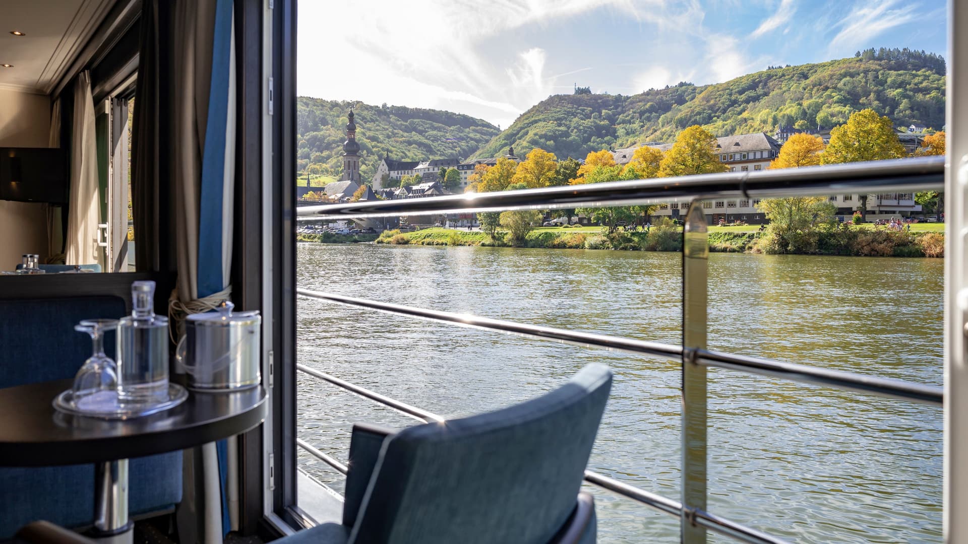 Open-air Panorama Suite aboard an Avalon Waterways ship featuring a wide sliding glass door framing a scenic river, a balcony railing, and a European hillside village with a church steeple.