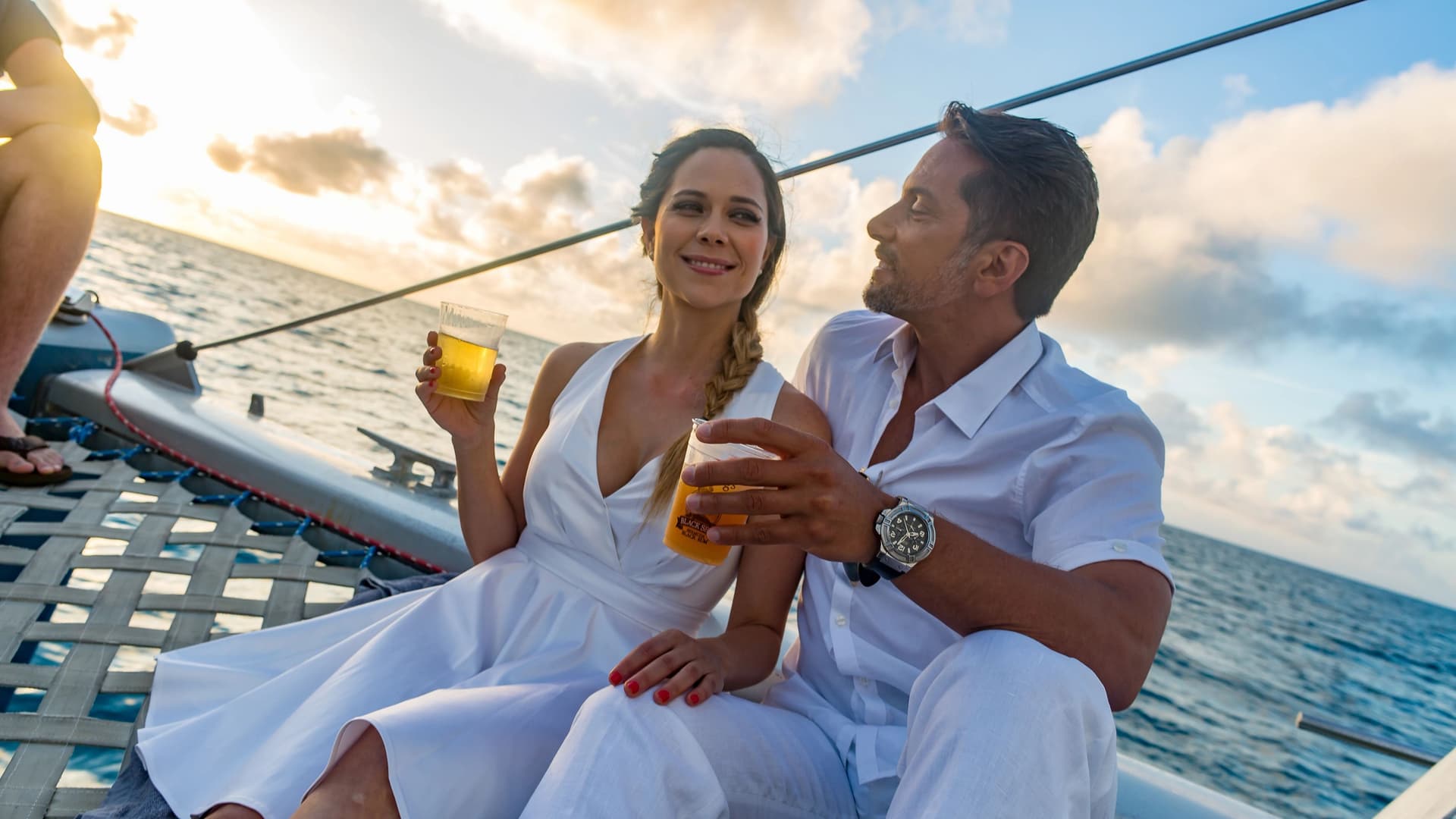 A man and a woman dressed in white clothes sit on the netting of a catamaran at sunset, holding drinks and looking lovingly at each other.