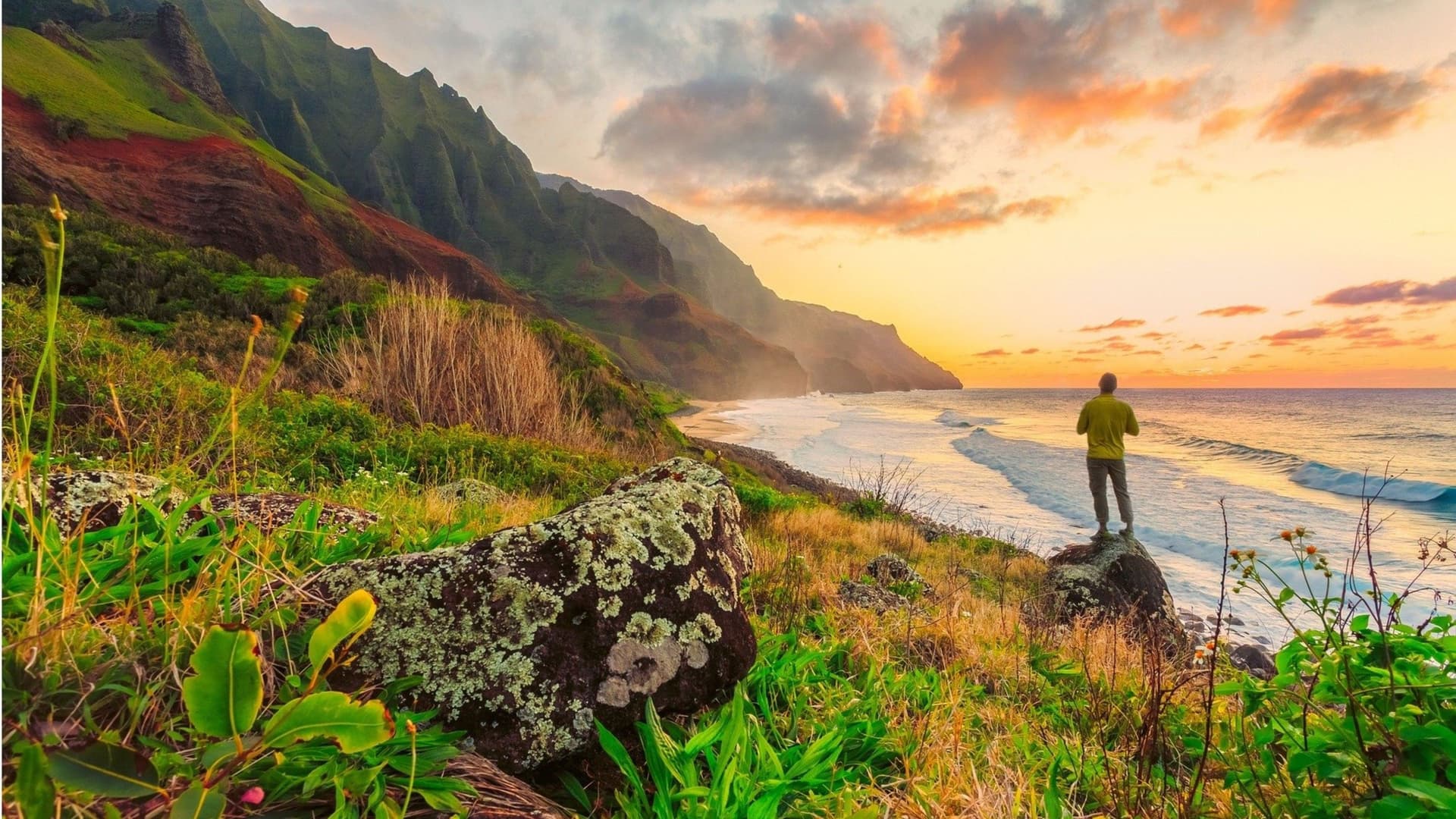 A hiker stands on a coastal cliff overlooking the Na Pali Coast's rugged mountains and glowing sunset.