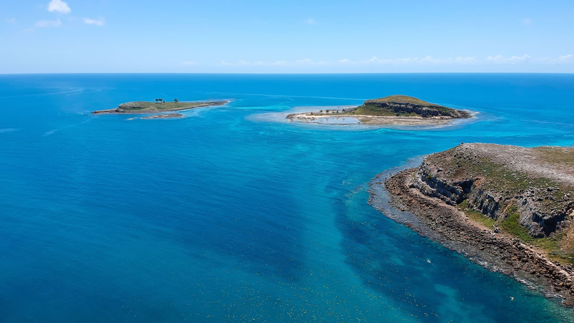 An aerial view of the Abrolhos Islands, with multiple small islands surrounded by crystal-clear turquoise water and reefs under a bright blue sky.